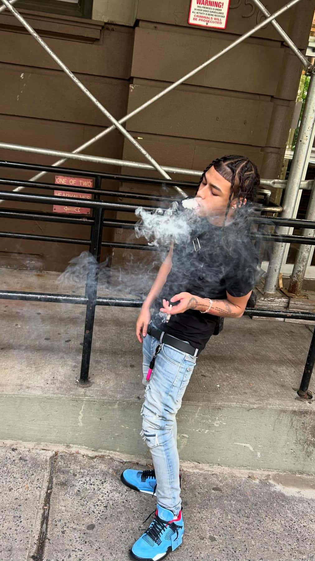  壁紙 — 一名穿著破洞牛仔褲和亮藍色運動鞋的年輕男子站在建築鷹架附近的城市人行道上，呼出濃厚的蒸氣。