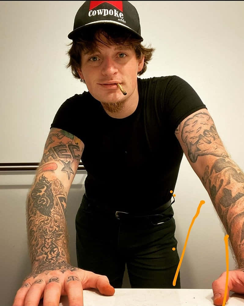  壁紙 — 一名刺青男子戴著黑色 'Cowpoke' 帽子和襯衫，嘴裡叼著香煙，靠在一面素牆上向前傾。