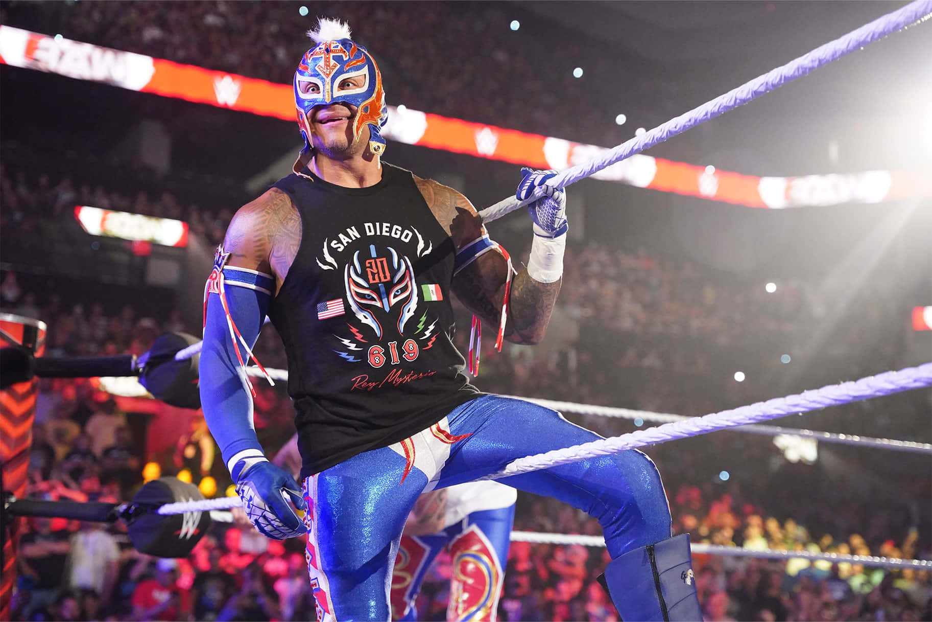  壁紙 — 一名戴著面具的藍橙色裝備 lucha libre 摔角手靠在燈光明亮的摔角場的繩索上，身後有歡呼的人群。