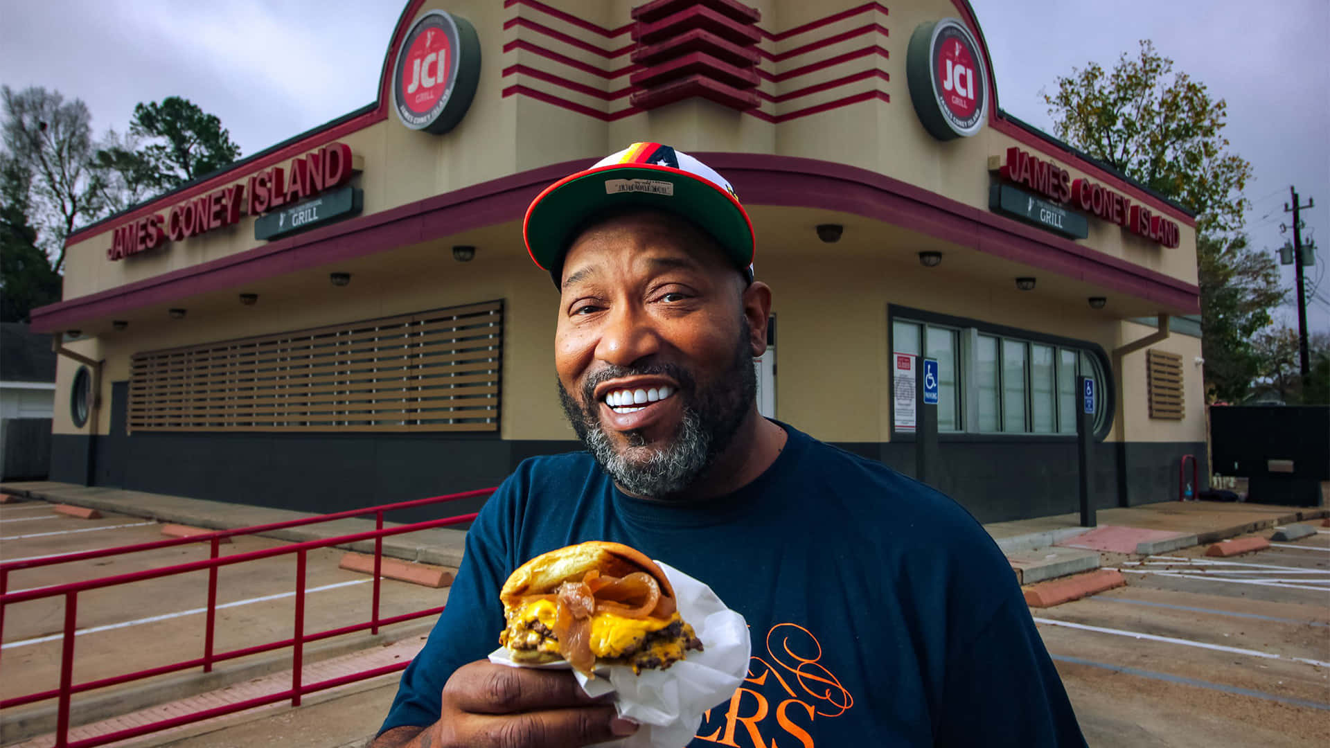  壁紙 — 一名戴帽子的微笑男子在標誌性的James Coney Island Grill外自豪地拿著一個餡料豐富的漢堡，餐廳有紅色欄杆和復古標牌。