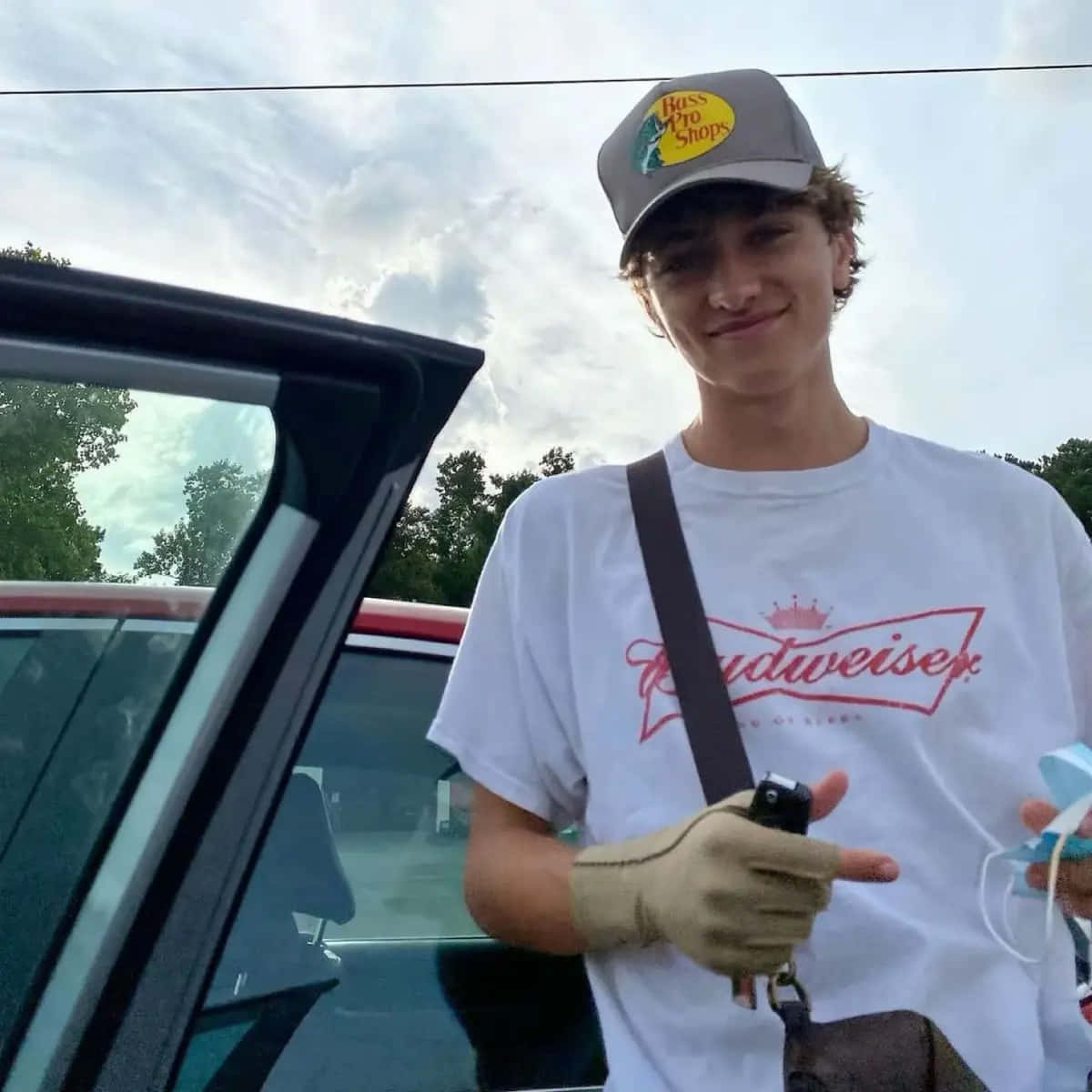  壁紙 — 一名戴著Bass Pro Shops帽子和穿著Budweiser T恤的年輕男子在多雲的天空下，在打開的車門旁微笑。