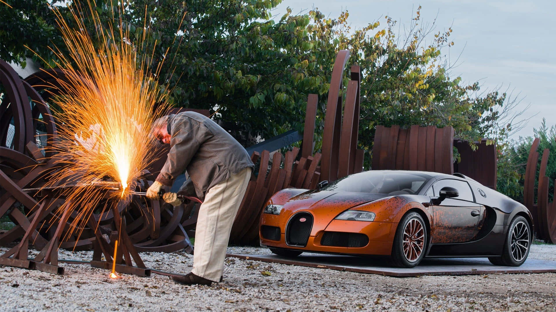 Bugatti Veyron 桌布 壁紙 — 一名焊接工在停泊於碎石上、靠近生鏽金屬雕塑的訂製橙色Bugatti Veyron旁邊強烈地產生火花。
