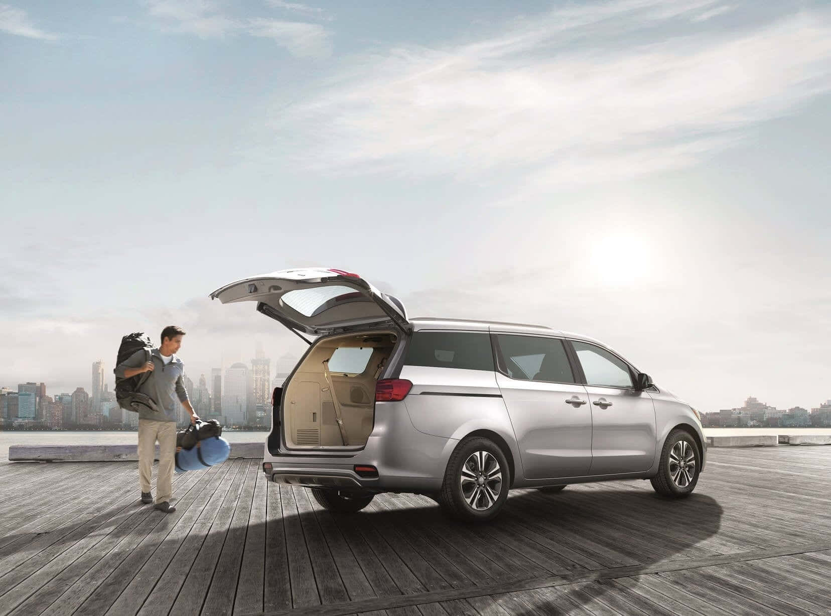 Kia Sedona 桌布 壁紙 — 一名男子帶著行李站在木製碼頭上，旁邊是一輛銀色廂型車，後門敞開，俯瞰著晴朗天空下的城市天際線。