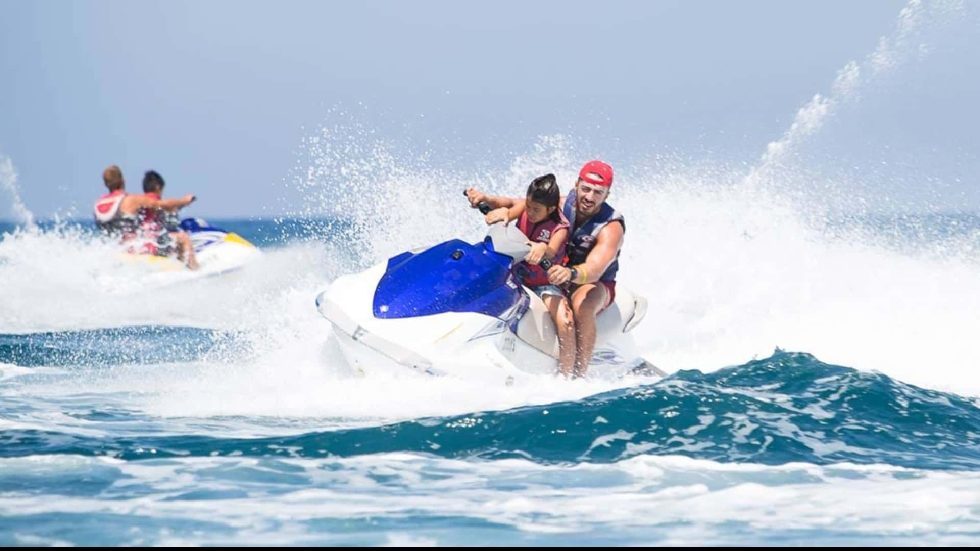 Beach Water Sports 桌布 壁紙 — 一名男子和一個小孩騎著藍白色的水上摩托車，在清澈藍天下穿越碧綠的海浪。