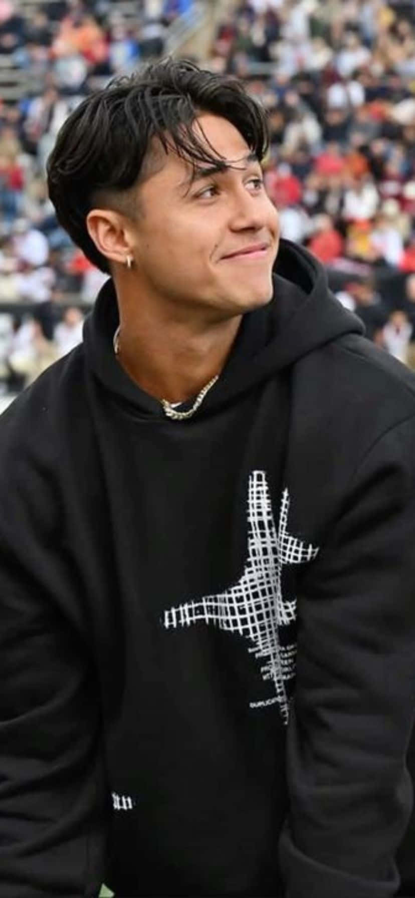  壁紙 — 一名身穿帶有白色圖案的黑色連帽衫的年輕男子微笑著仰望擠滿人的體育場。