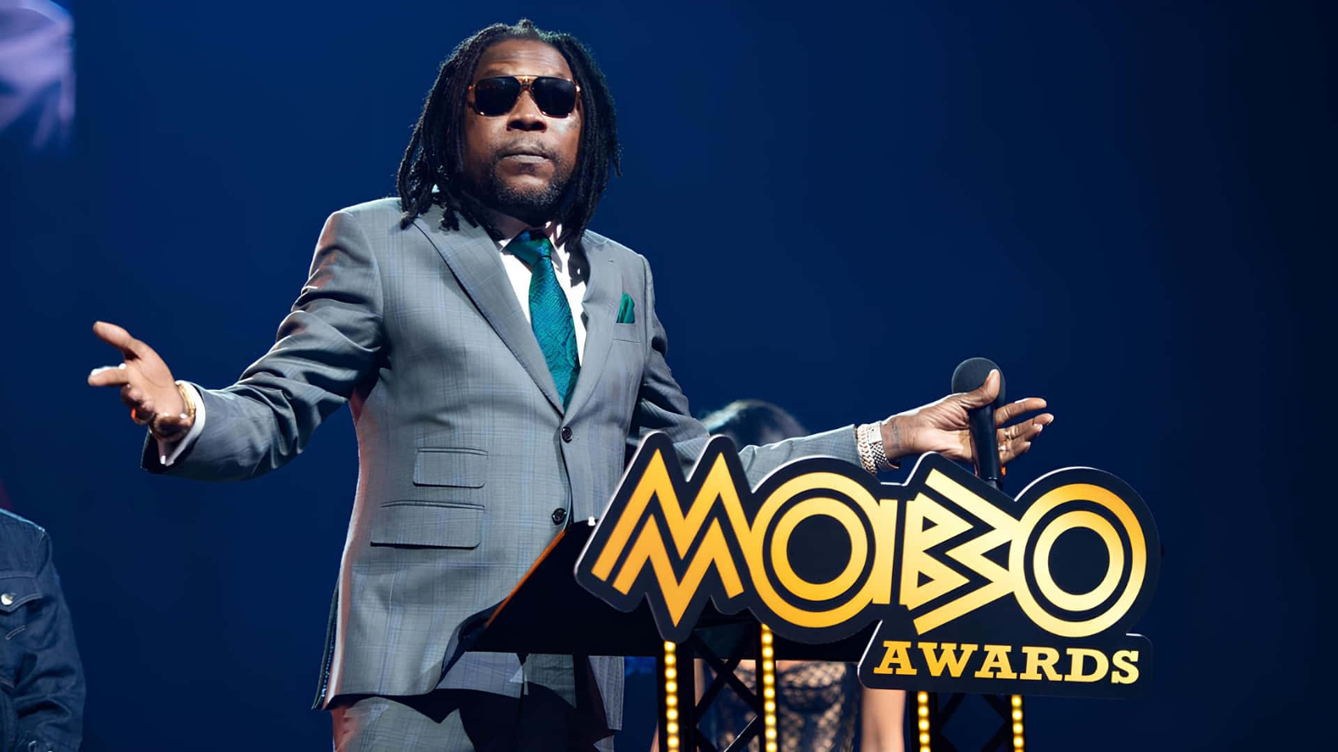  壁紙 — 一名身穿灰色西裝和太陽眼鏡的男子站在MOBO Awards頒獎台上，在藍色舞台燈光下拿著麥克風。