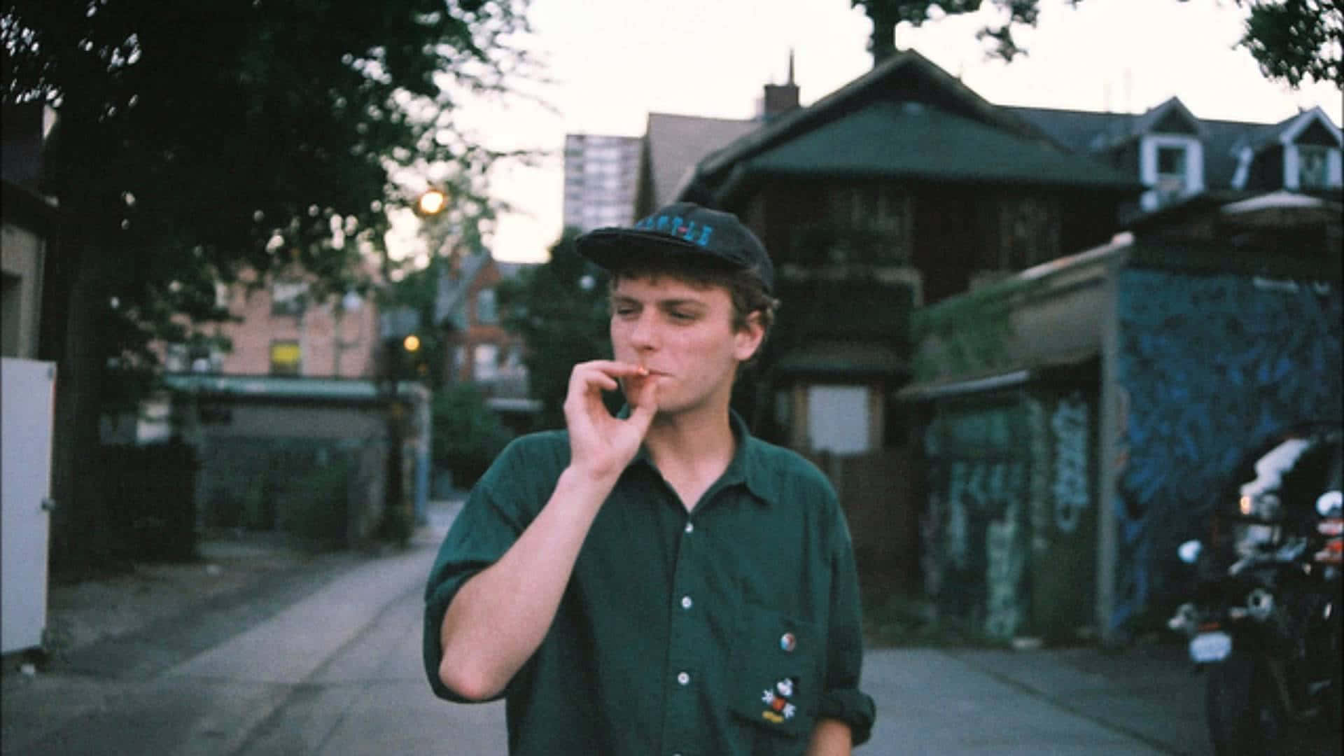  壁紙 — 一名身穿綠色襯衫和帽子的年輕男子在黃昏時分於佈滿塗鴉的城市街道上吸煙。