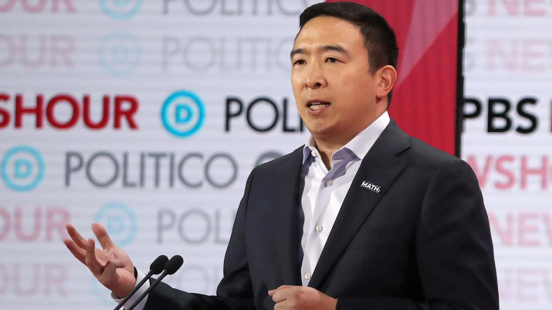 美國 桌布 壁紙 — 一名身穿深色西裝的男子在 Politico 和 PBS 標誌的背景前，在講台上說話時做手勢。