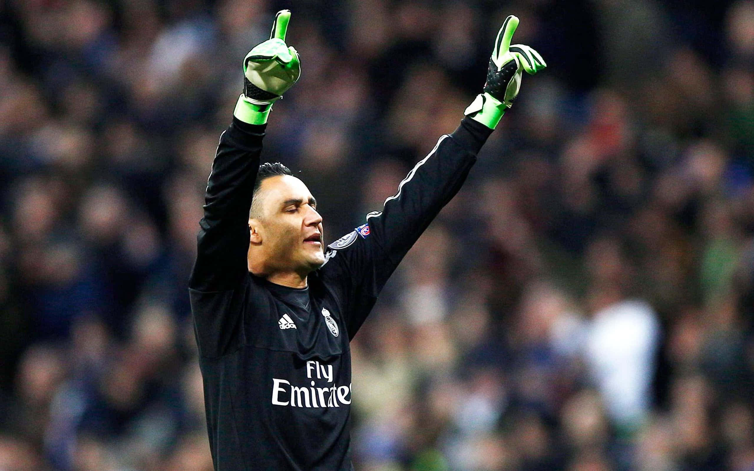 Keylor Navas 桌布 壁紙 — 一名身穿黑色球衣的足球守門員，在模糊的體育場人群中高舉戴著手套的雙手慶祝勝利。