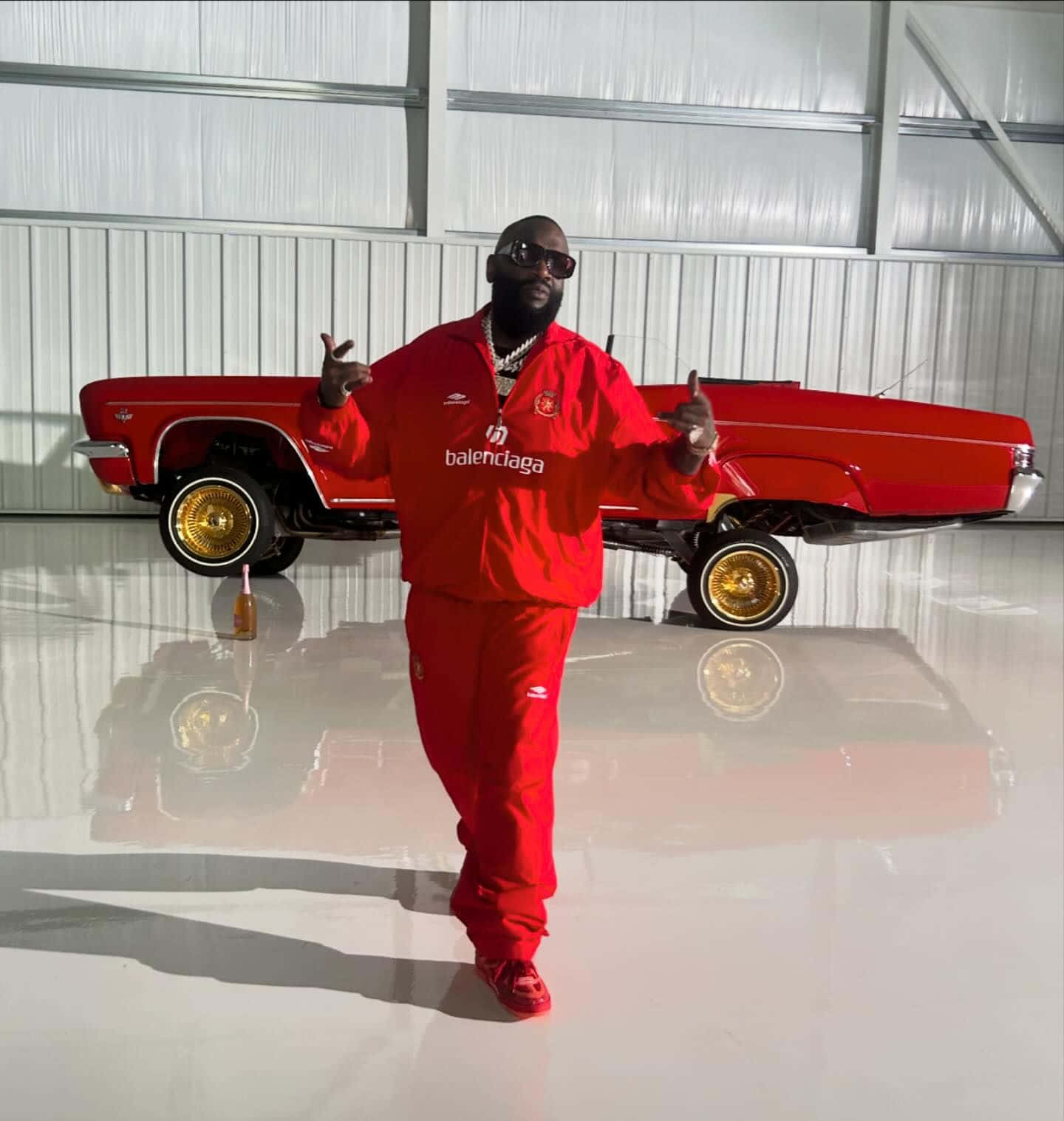  壁紙 — 一名身穿紅色Balenciaga運動服的男子在明亮的金屬車庫內，自信地站在一輛配有金色輪圈的紅色經典汽車前擺姿勢。