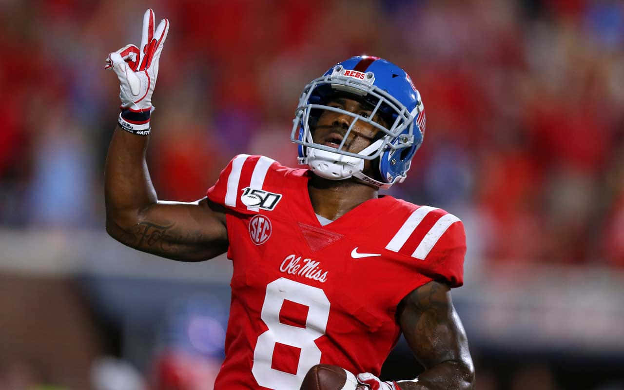  壁紙 — 一名身穿紅色Ole Miss 8號球衣的美式足球員在球場燈光下舉起球並做出和平手勢慶祝。