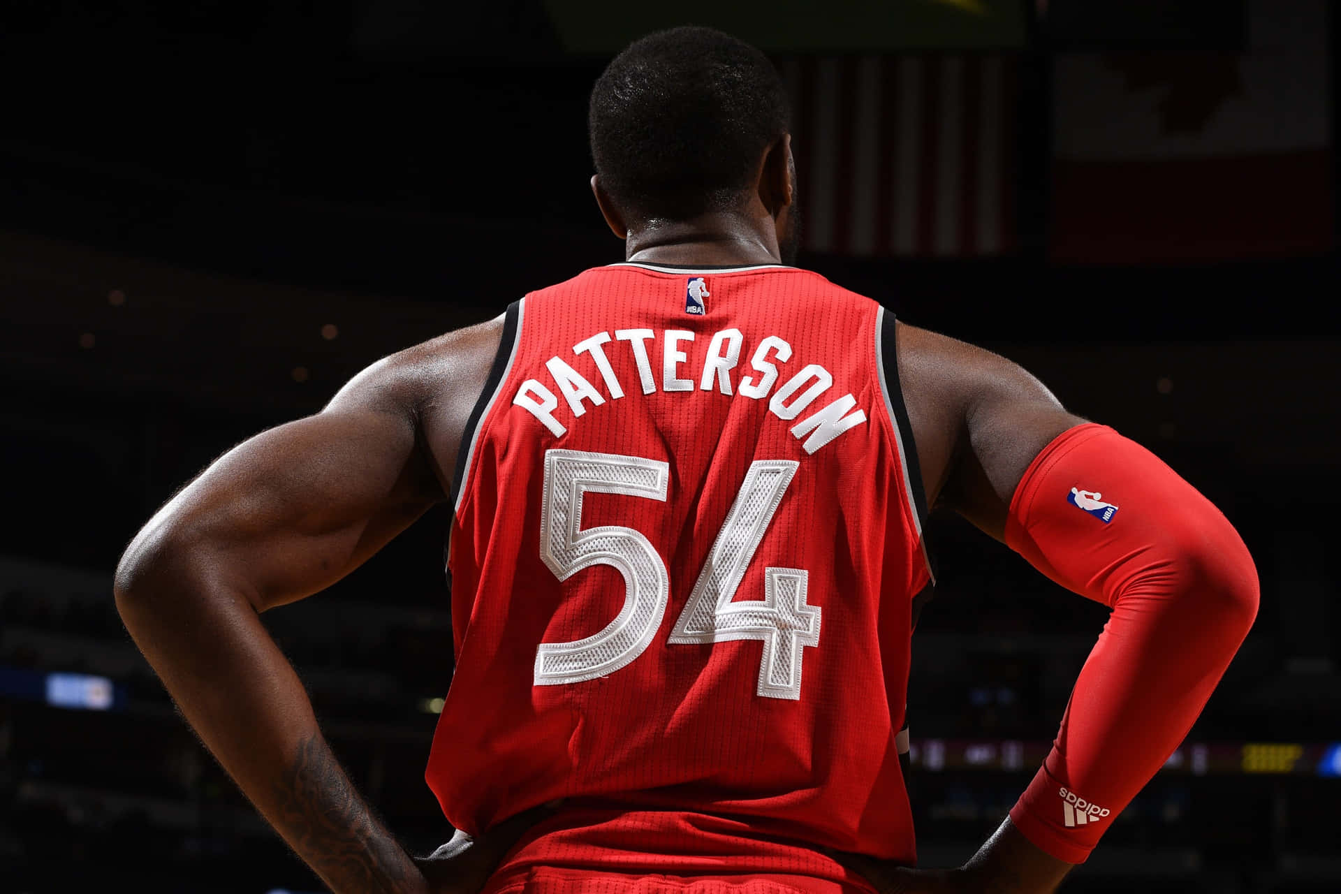  壁紙 — 一名身穿紅色Patterson 54球衣的籃球運動員在燈光昏暗的球場上背對鏡頭，雙手叉腰站立。