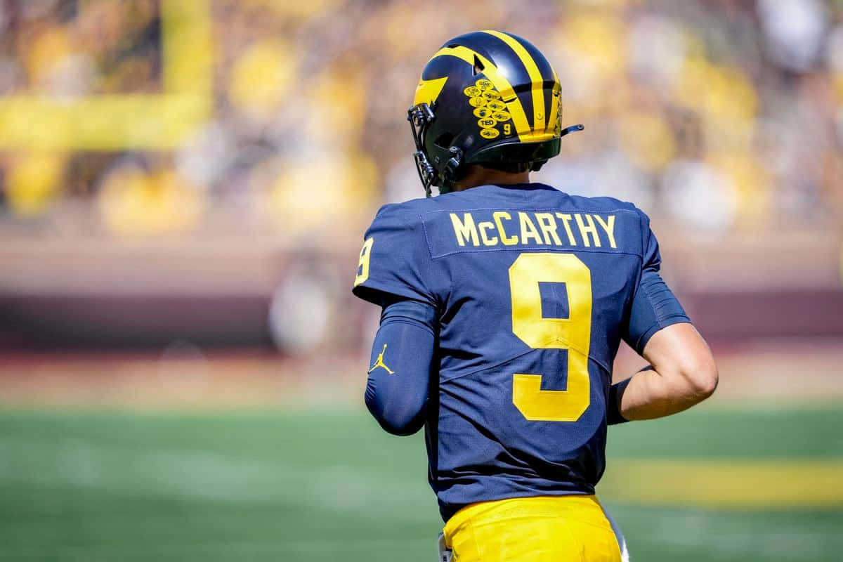  壁紙 — 一名身穿藍黃色球衣、背上印有McCarthy名字和9號的足球員站在球場上，背景是模糊的體育場座位。