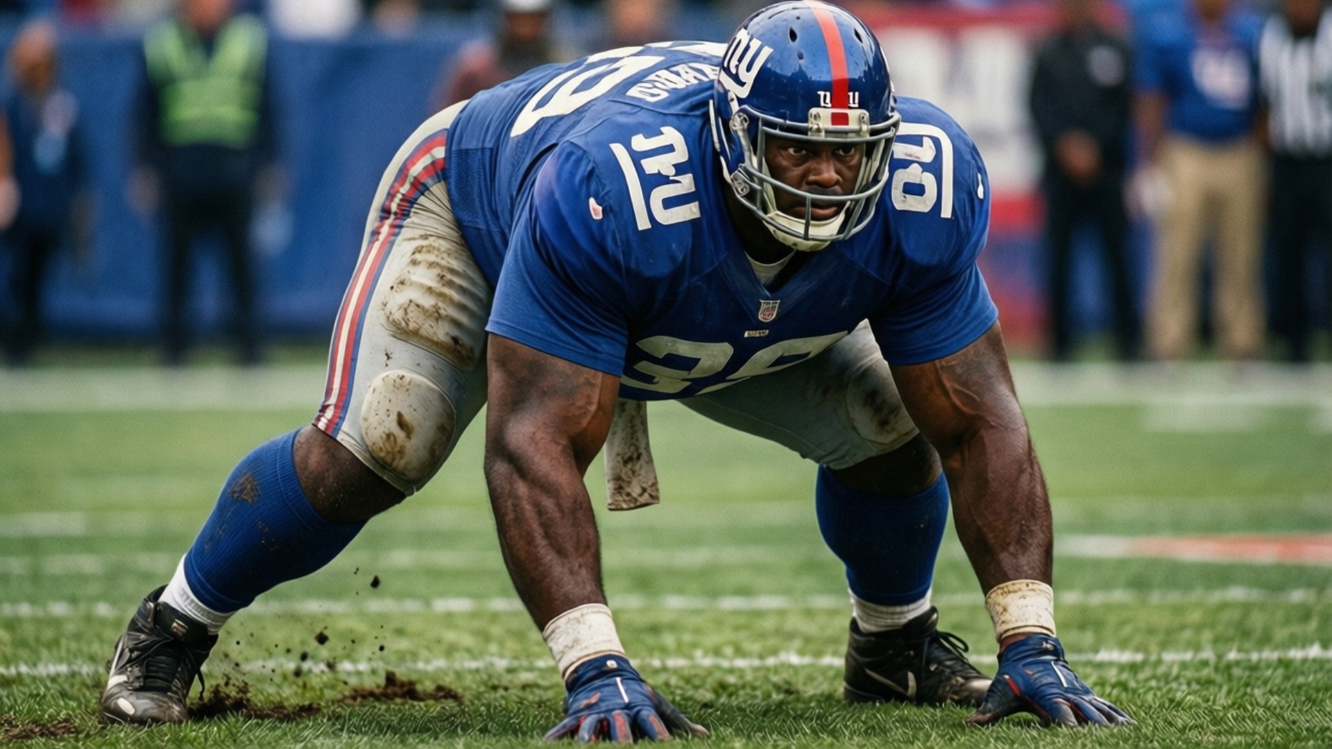 Giants Defensive Stance 壁紙 — 一名身穿藍色球衣的紐約巨人隊美式足球員在泥濘的綠色草地上低蹲，泥土從他的鞋釘上飛濺。