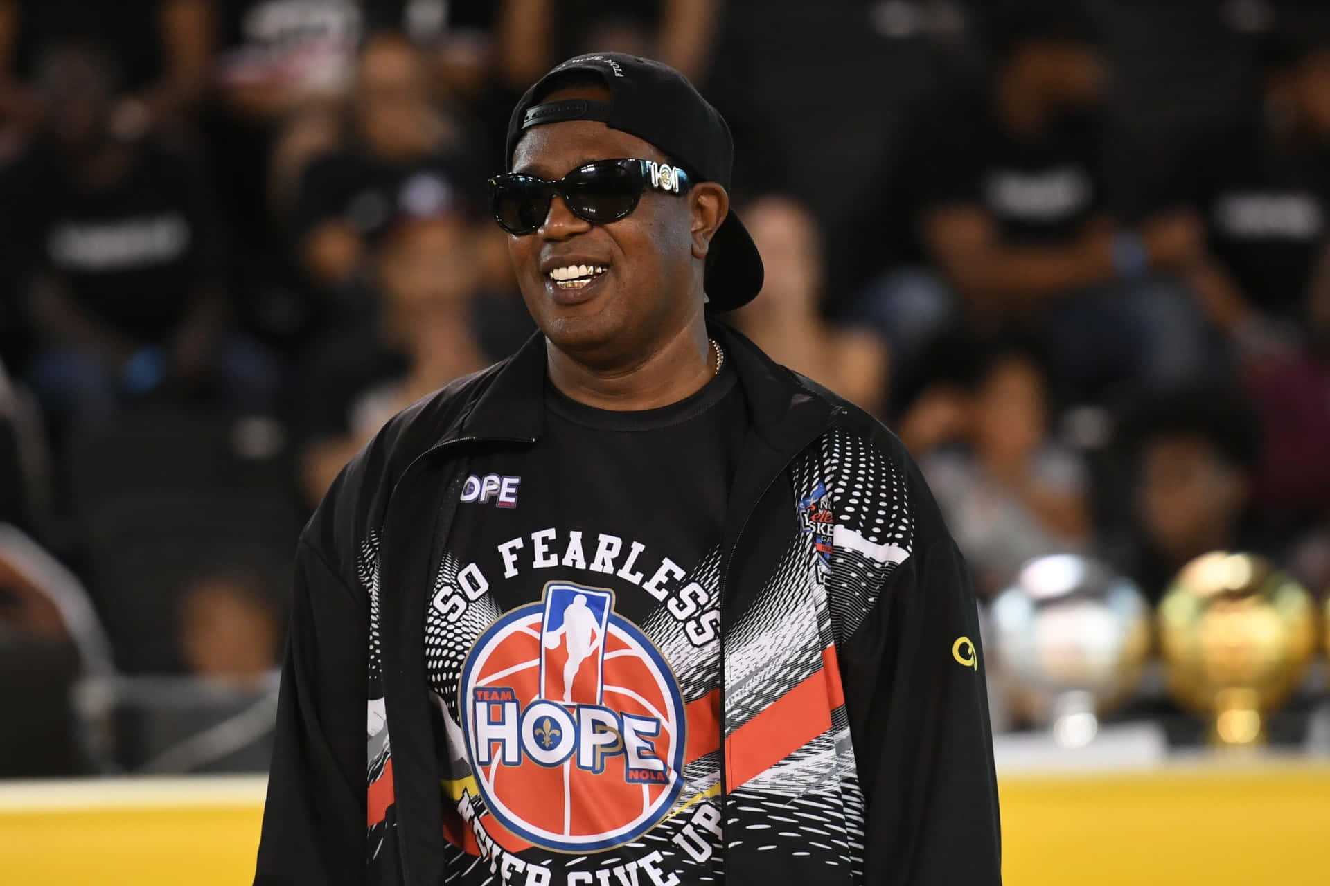 壁紙 — 一名微笑的男子身穿黑色「So Fearless Hope」NBA外套和帽子，站在擠滿人的場館內，身後可見獎盃。