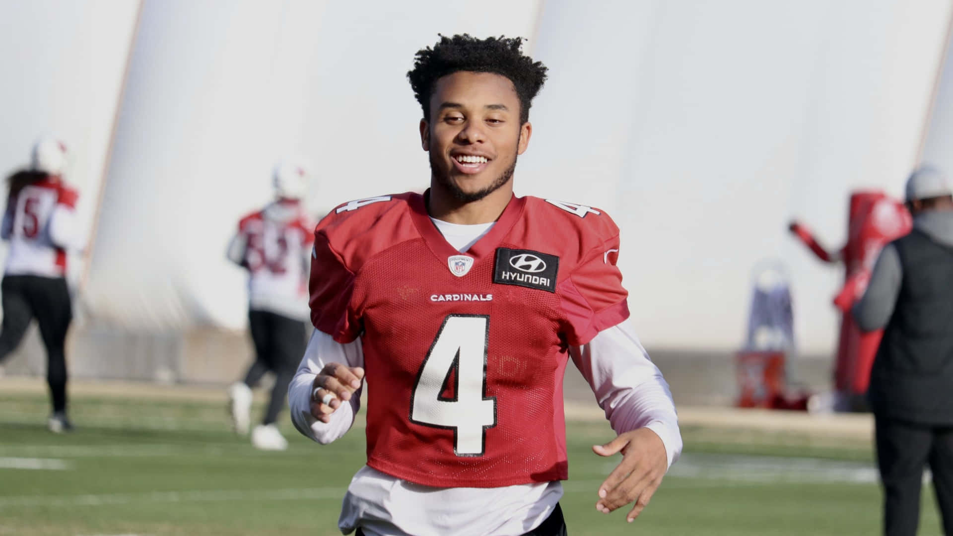  壁紙 — 一名微笑的美式足球員，身穿紅色 Cardinals 球衣，背號 4，在陽光明媚的訓練場上全力衝刺。