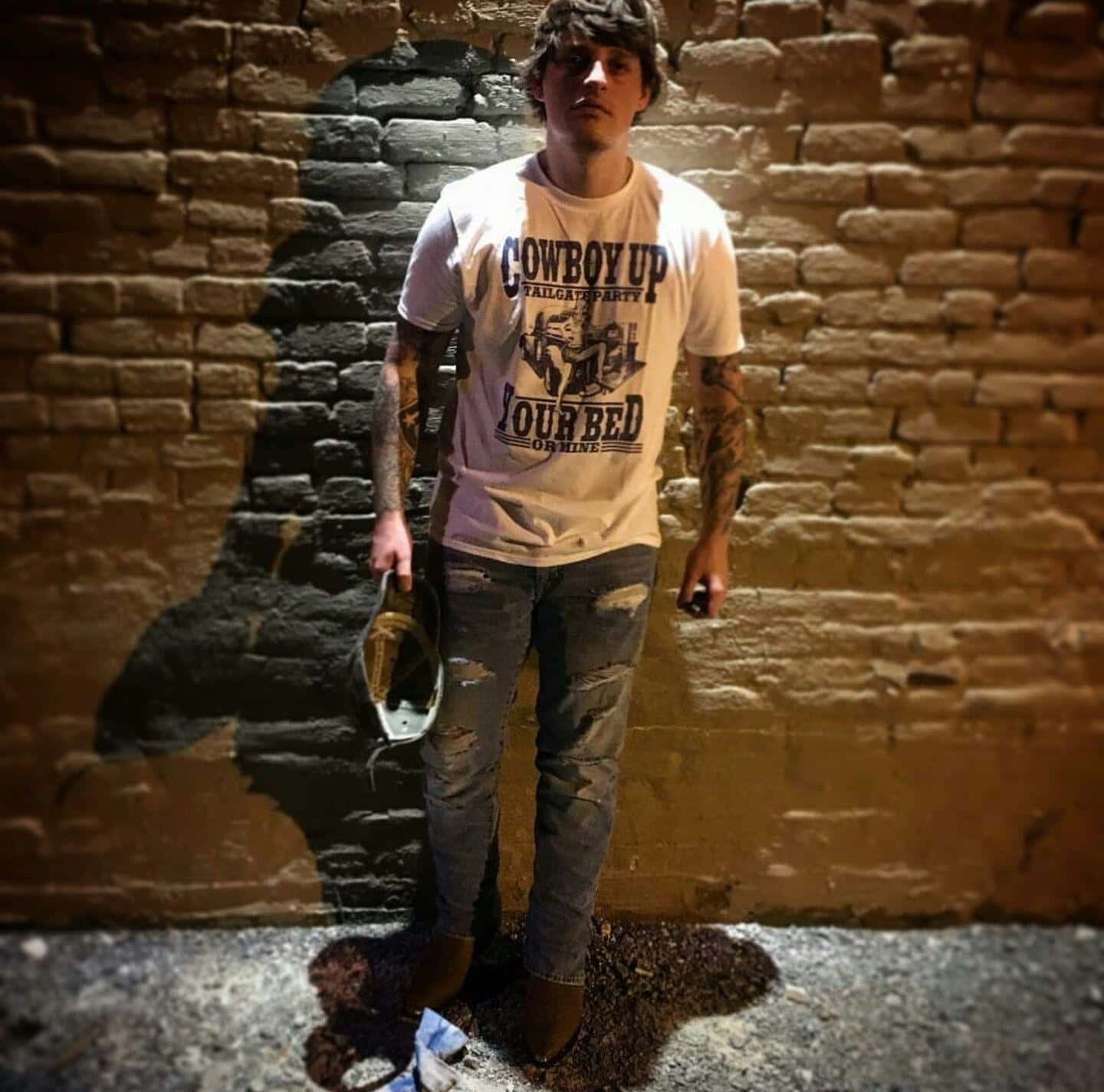  壁紙 — 一名有刺青的年輕男子在夜間靠著磚牆，在溫暖的人造燈光下拿著帽子和手機。