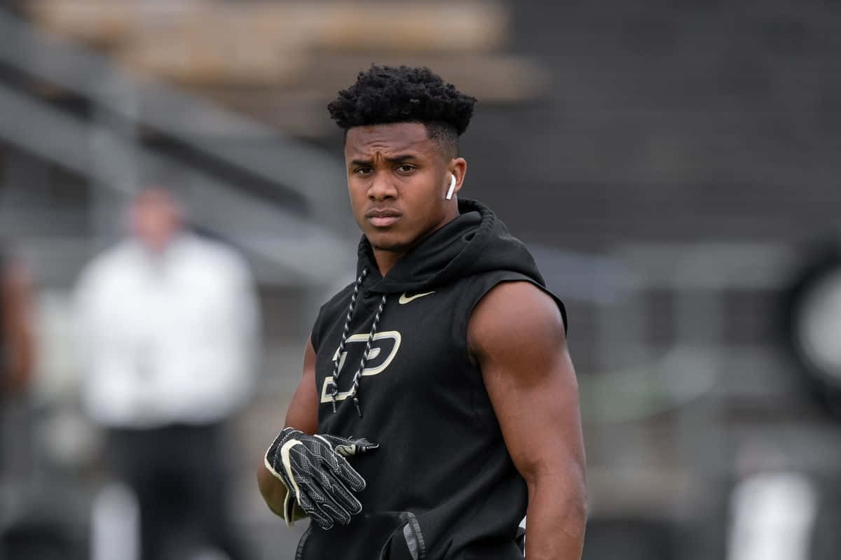 壁紙 — 一名專注的美式足球員身穿黑色裝備，戴著白色耳塞，在陰天下練習時站在球場上。