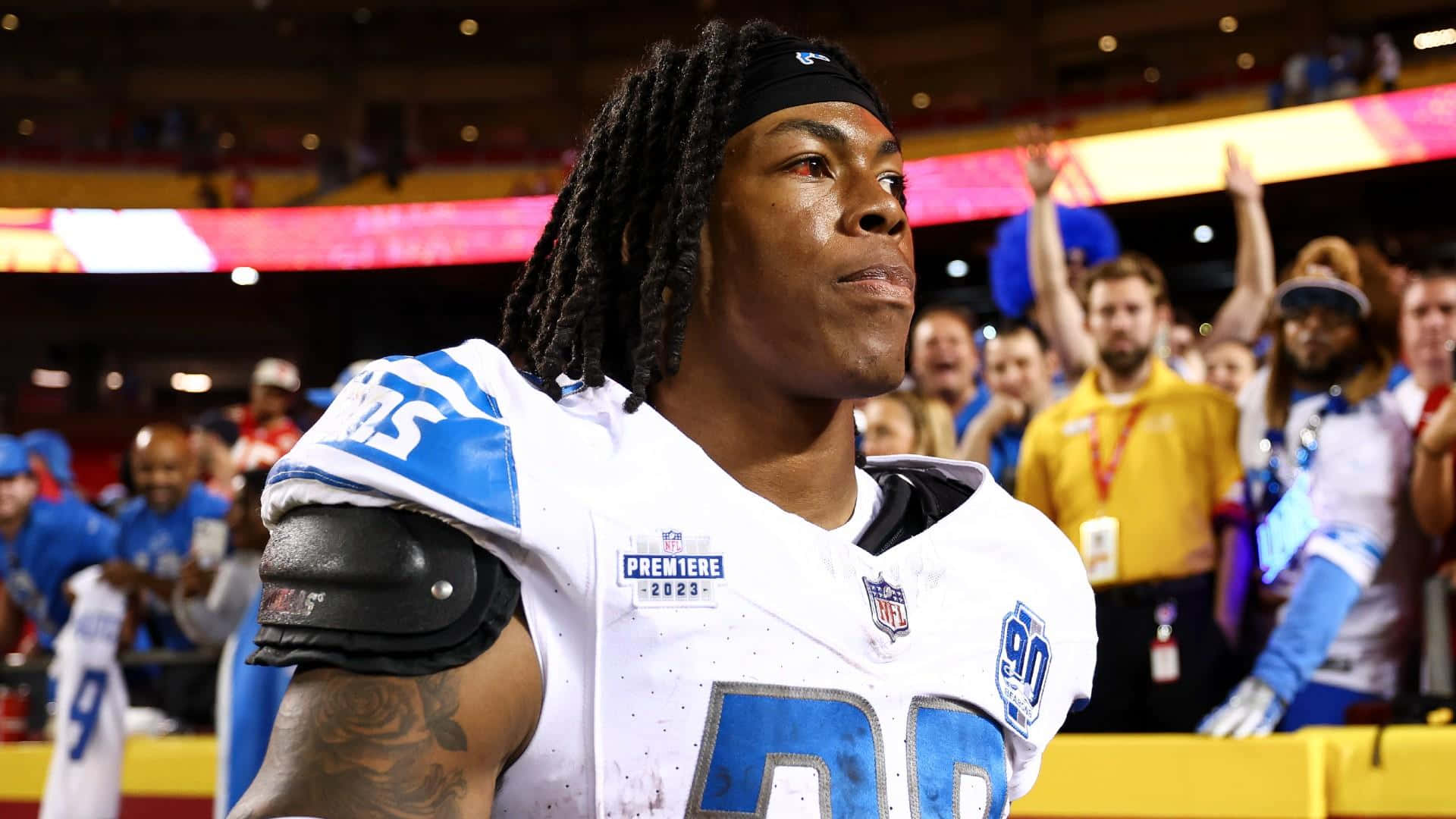  壁紙 — 一名專注的NFL美式足球員身穿白色和藍色球衣，站在球場上，背景有體育場燈光和歡呼的球迷。