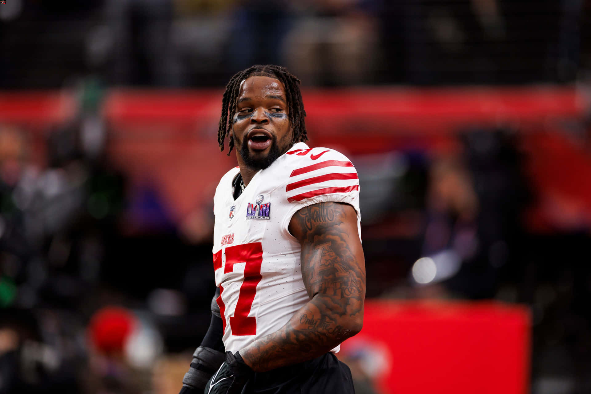 壁紙 — 一名專注的NFL球員身穿白色球衣，紅色27號碼和黑色手套，站在球場上，手臂上的刺青清晰可見。