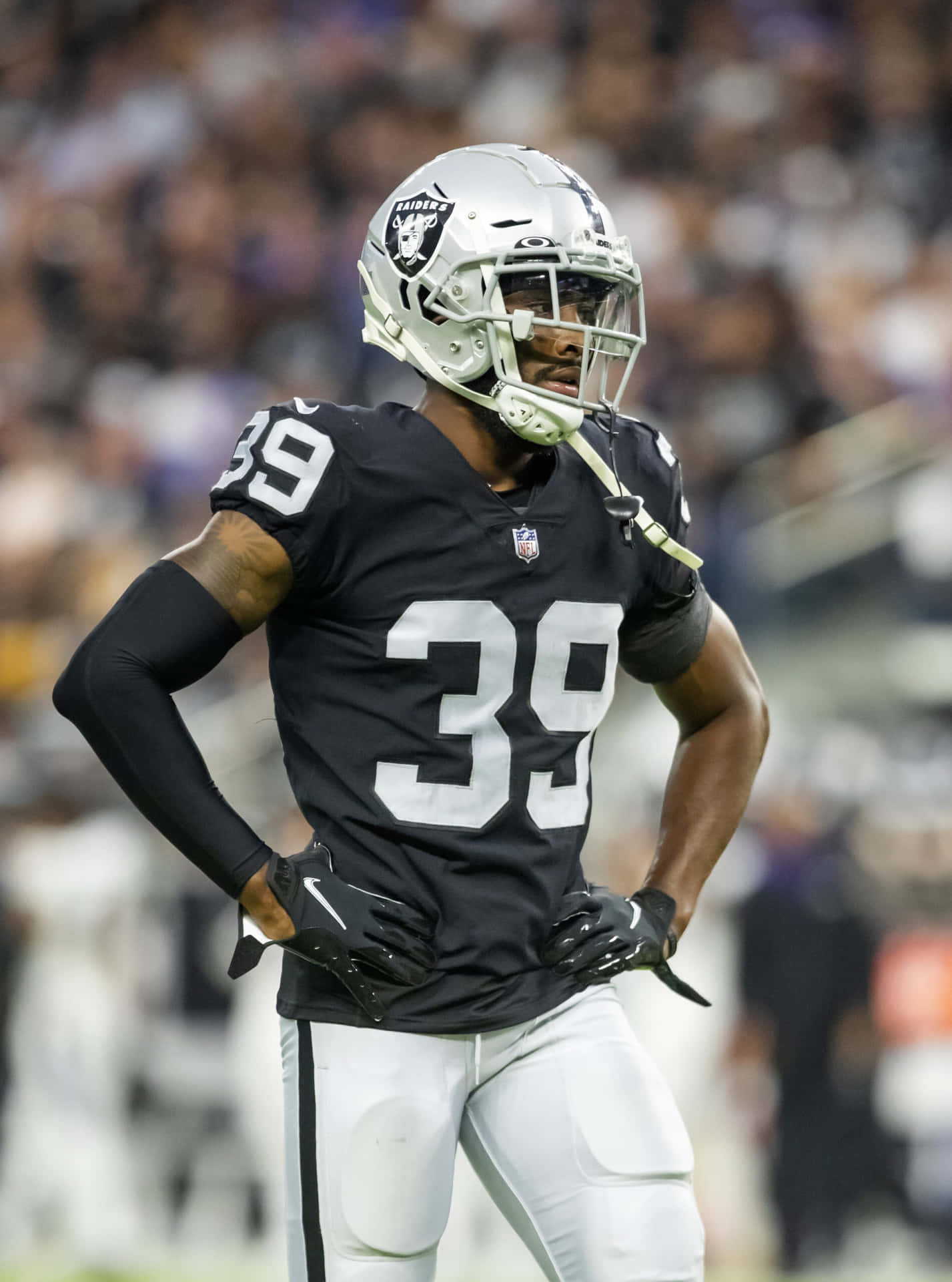  壁紙 — 一名專注的NFL球員身穿黑色和銀色的Raiders球衣，雙手叉腰站在燈光下的足球場上。