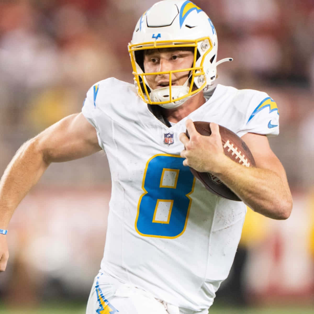  壁紙 — 一名專注的NFL四分衛身穿白色和藍色制服，在體育館燈光下比賽中持球衝刺。