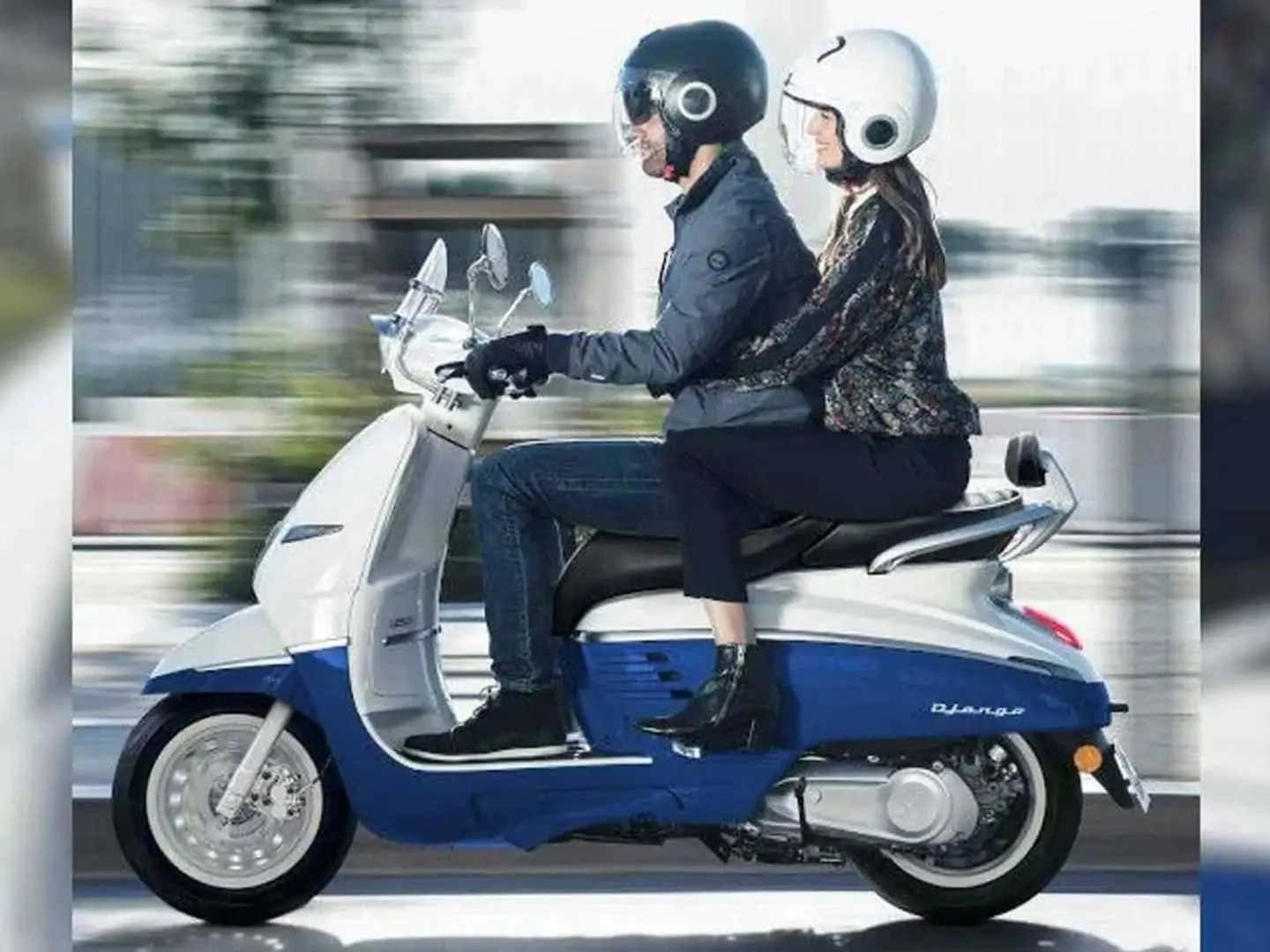 Peugeot Motorcycle 桌布 壁紙 — 一男一女戴著頭盔，騎著藍白相間的機車行駛在模糊的城市街道上。