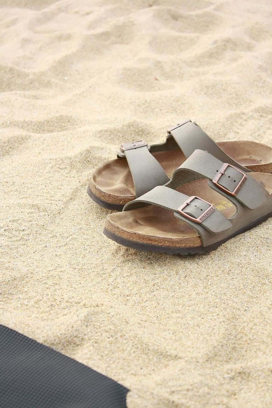  壁紙 — 一雙灰色Birkenstock涼鞋放置在細沙上，附近有深色毛巾角。