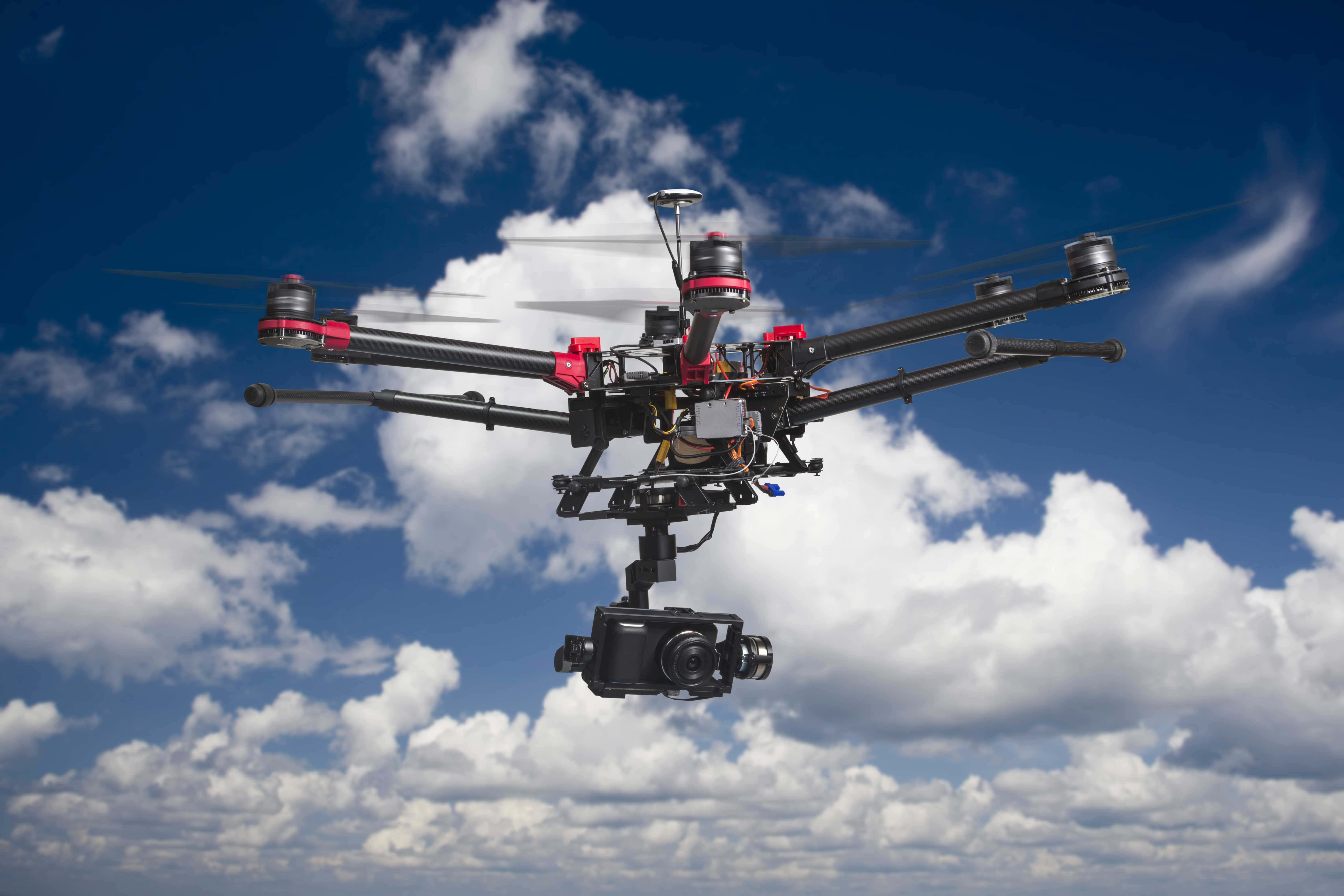 Drone Camera 桌布 壁紙 — 一台黑色和紅色的四軸無人機，帶有安裝的攝影機，在充滿蓬鬆白雲的鮮豔藍天中盤旋。