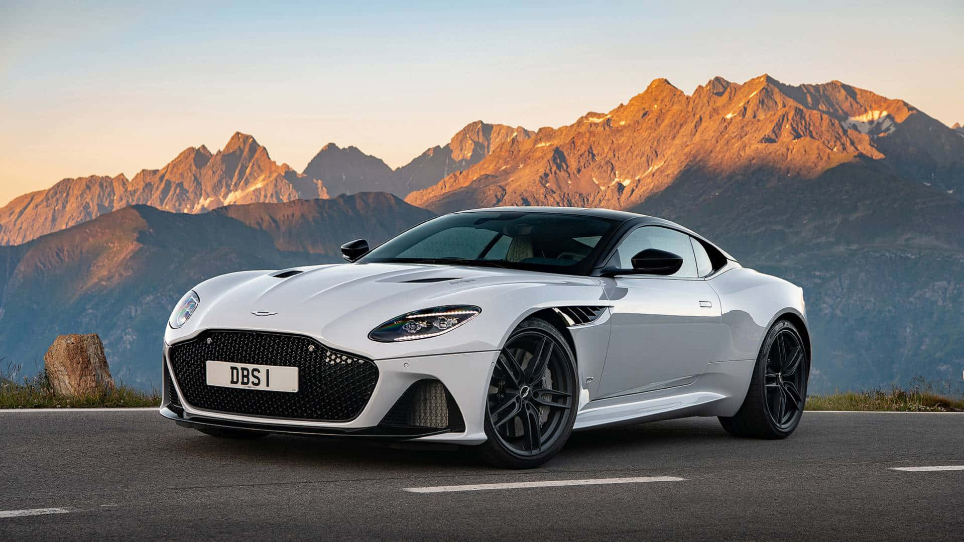 Aston Martin Dbs Superleggera 桌布 壁紙 — 一台流線型白色Aston Martin DBS I跑車停在山路上，背景是日落時分金色的山峰。