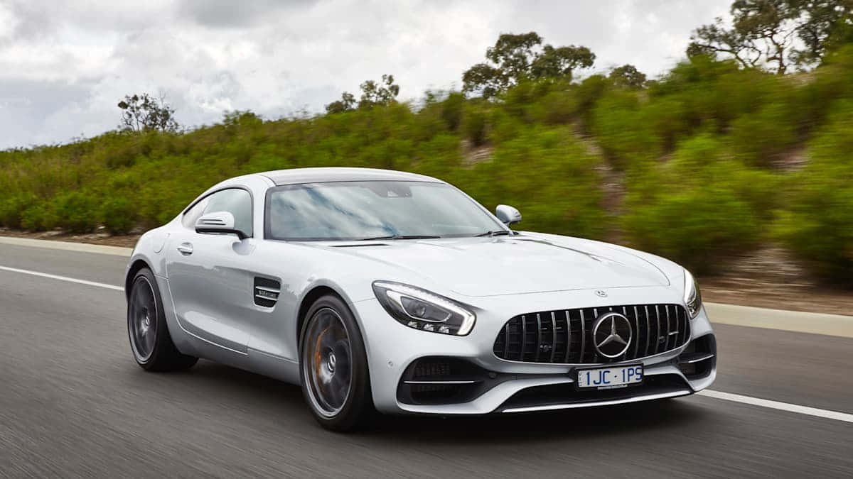 Mercedes Benz Amg Gt 桌布 壁紙 — 一台流線型白色 Mercedes-AMG GT 跑車在陰天之下，沿著蜿蜒的道路穿過翠綠的山丘飛馳。