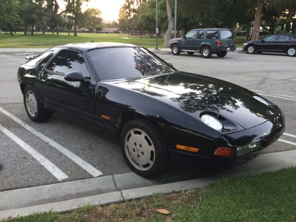 Porsche 928 桌布 壁紙 — 一台流線型的黑色保時捷928在停車場閃閃發光，黃金時段的光線反射在其拋光表面上。