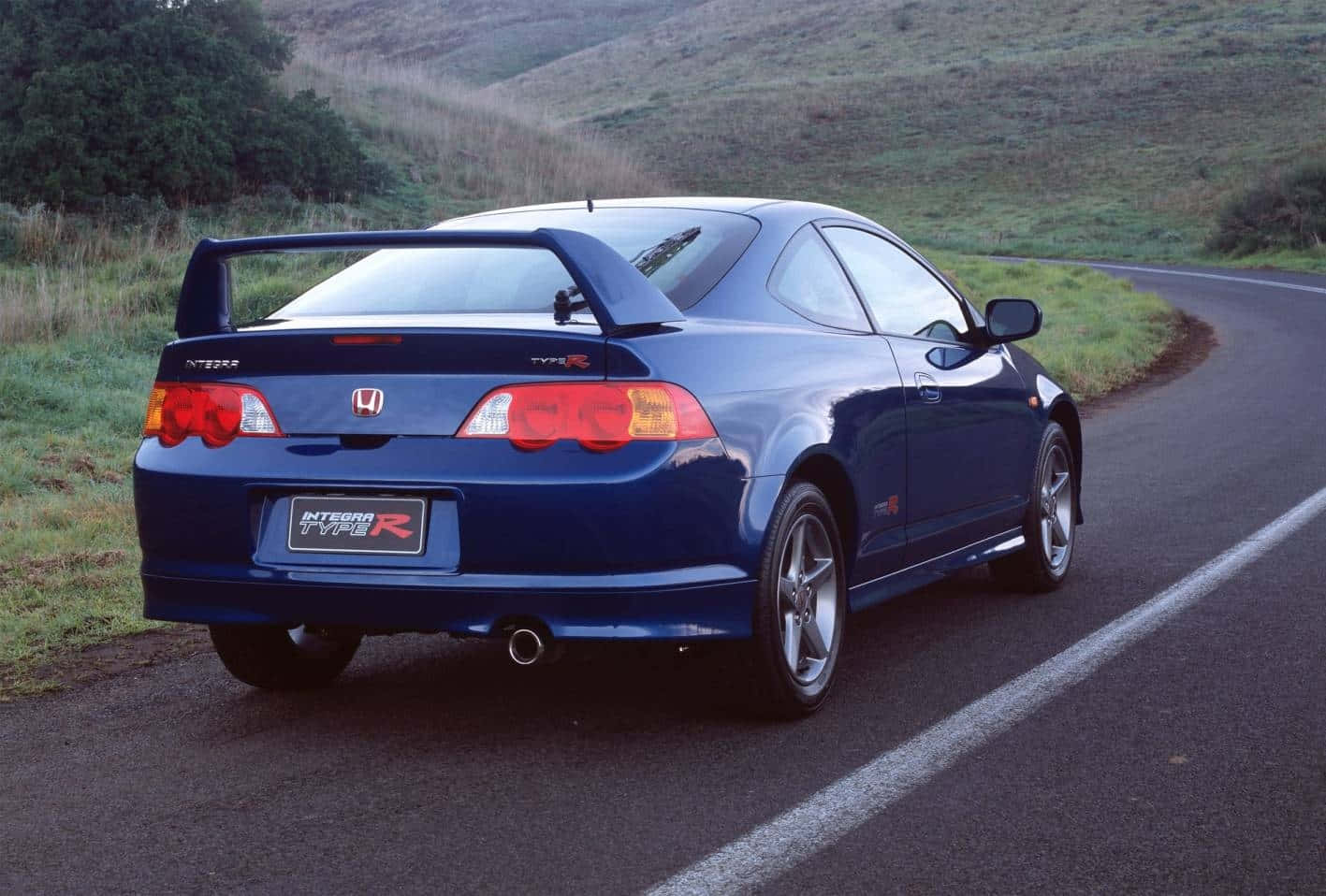 Honda Integra 桌布 壁紙 — 一台流線型的藍色本田Integra Type R轎跑車沿著一條蜿蜒的鄉間道路行駛，兩旁是長滿青草的山丘。