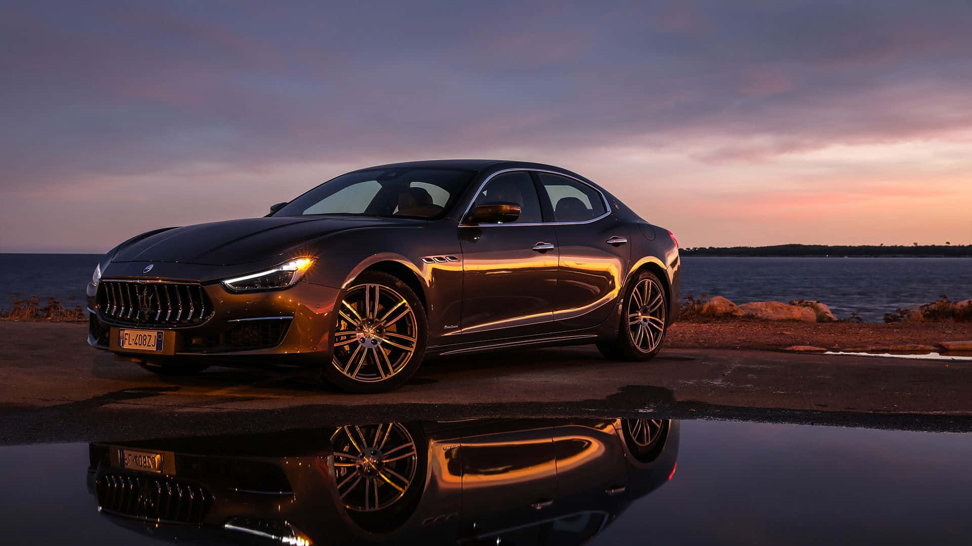 Maserati Ghibli 桌布 壁紙 — 一台流線型黑色瑪莎拉蒂轎車在黃昏閃耀，倒影映照在海邊道路旁的水坑中。