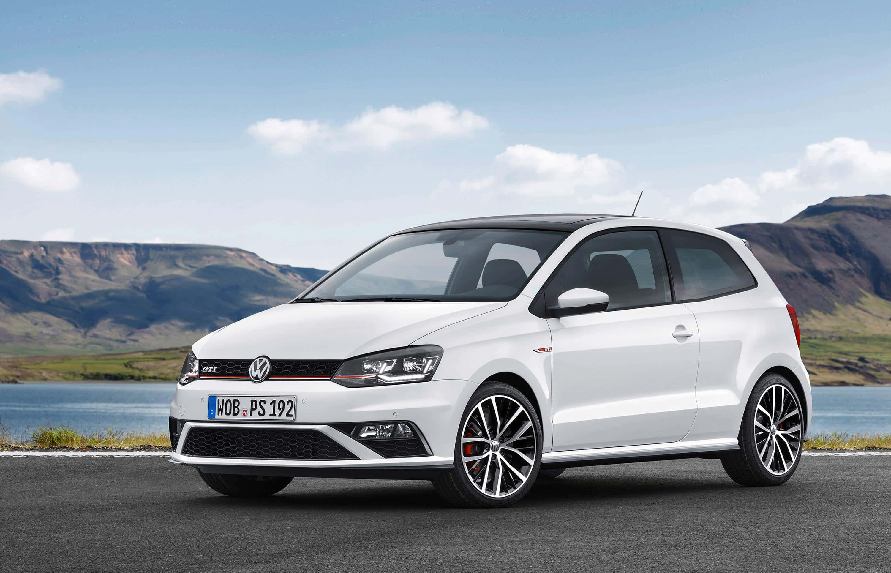 Volkswagen Polo 桌布 壁紙 — 一台時尚的白色福斯Polo GTI停泊在湖邊的道路上，背景是起伏的丘陵，天空部分多雲。