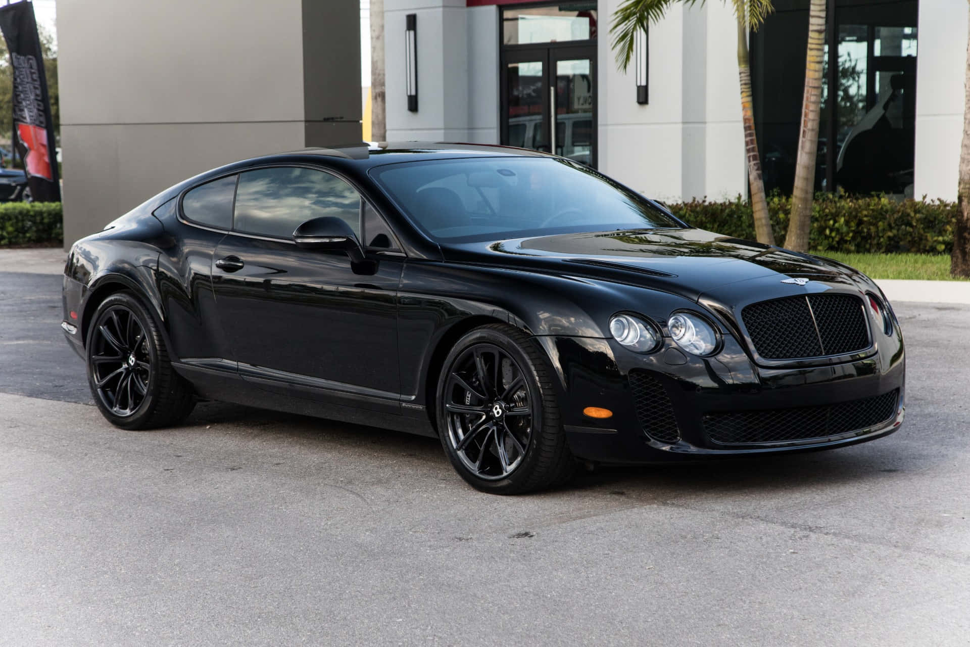Bentley Continental Supersports 桌布 壁紙 — 一台時尚的黑色賓利 Continental GT 雙門轎跑車在日光下閃閃發光，停放在鋪砌的停車場上，旁邊是現代建築和棕櫚樹。