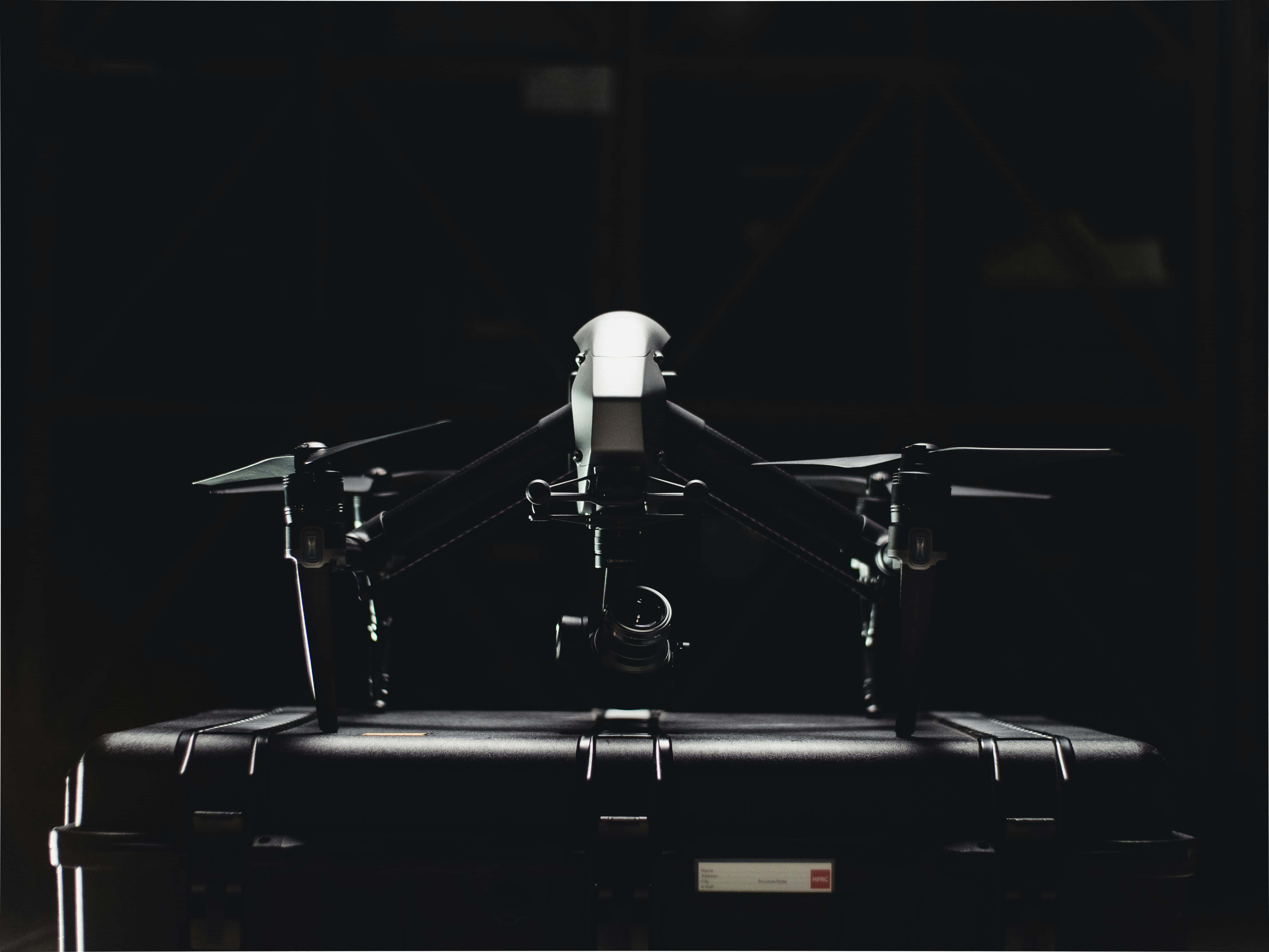 Drone Camera 桌布 壁紙 — 一台時尚的黑色無人機棲息在堅固的保護殼上，處於昏暗的工業空間，帶有戲劇性的背光效果。