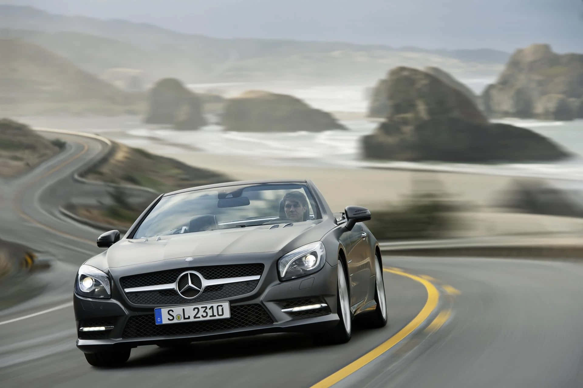 Mercedes Benz Sl-class 桌布 壁紙 — 一台時尚的灰色梅賽德斯敞篷車沿著海岸公路飛馳，背景是模糊的海洋和懸崖。
