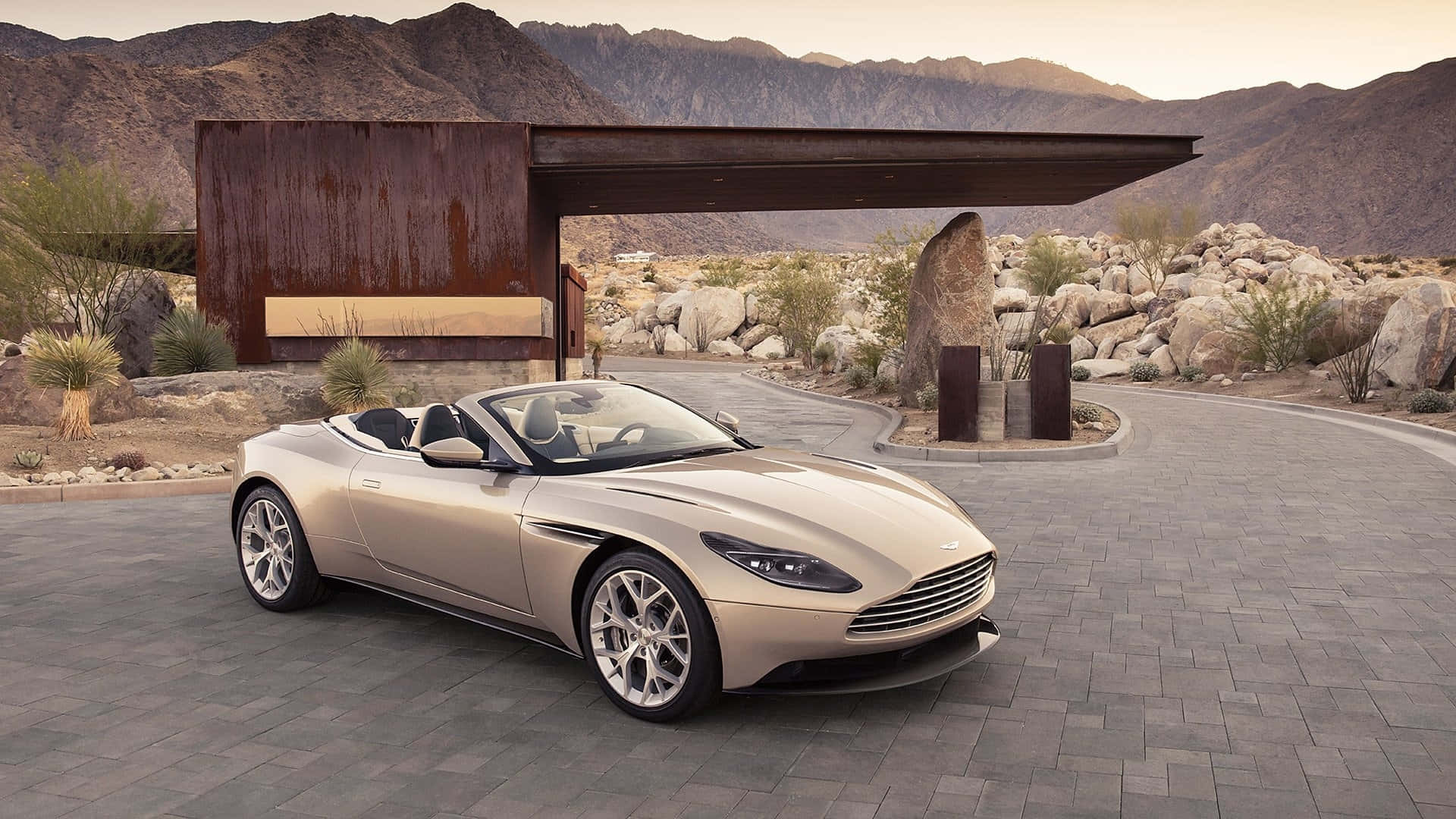 Aston Martin Db11 桌布 壁紙 — 一台時尚的米色Aston Martin DB9敞篷車停在石頭車道上，旁邊是現代鏽蝕金屬結構，背景有山脈。