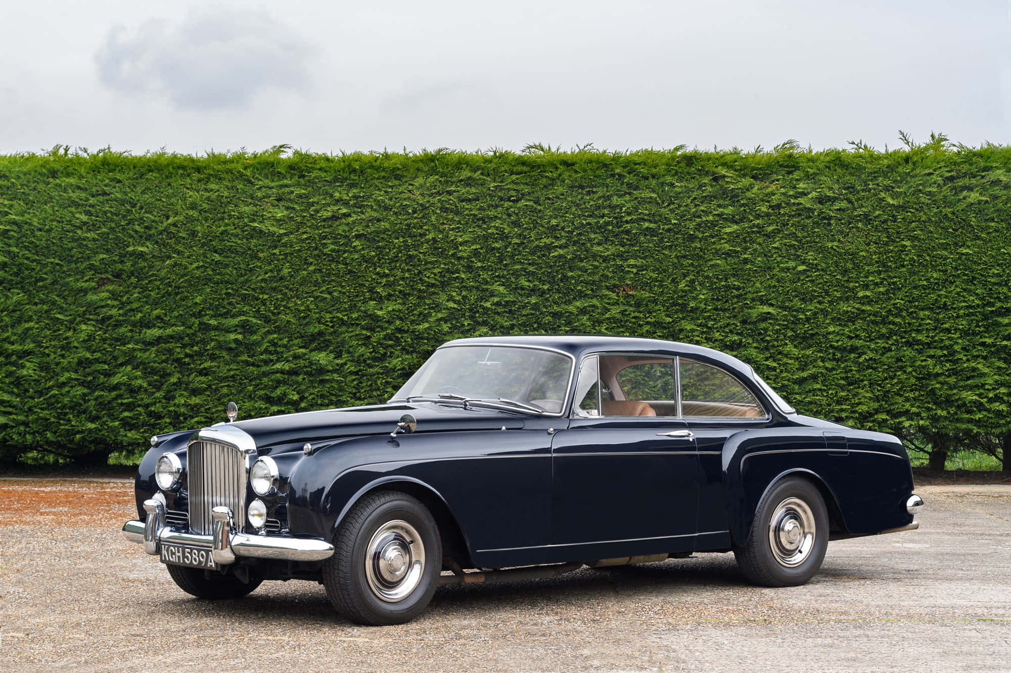 Bentley S2 桌布 壁紙 — 一台時尚的深藍色經典汽車，帶有鍍鉻裝飾，停泊在碎石路上，前面是高大的綠色樹籬，天空多雲。