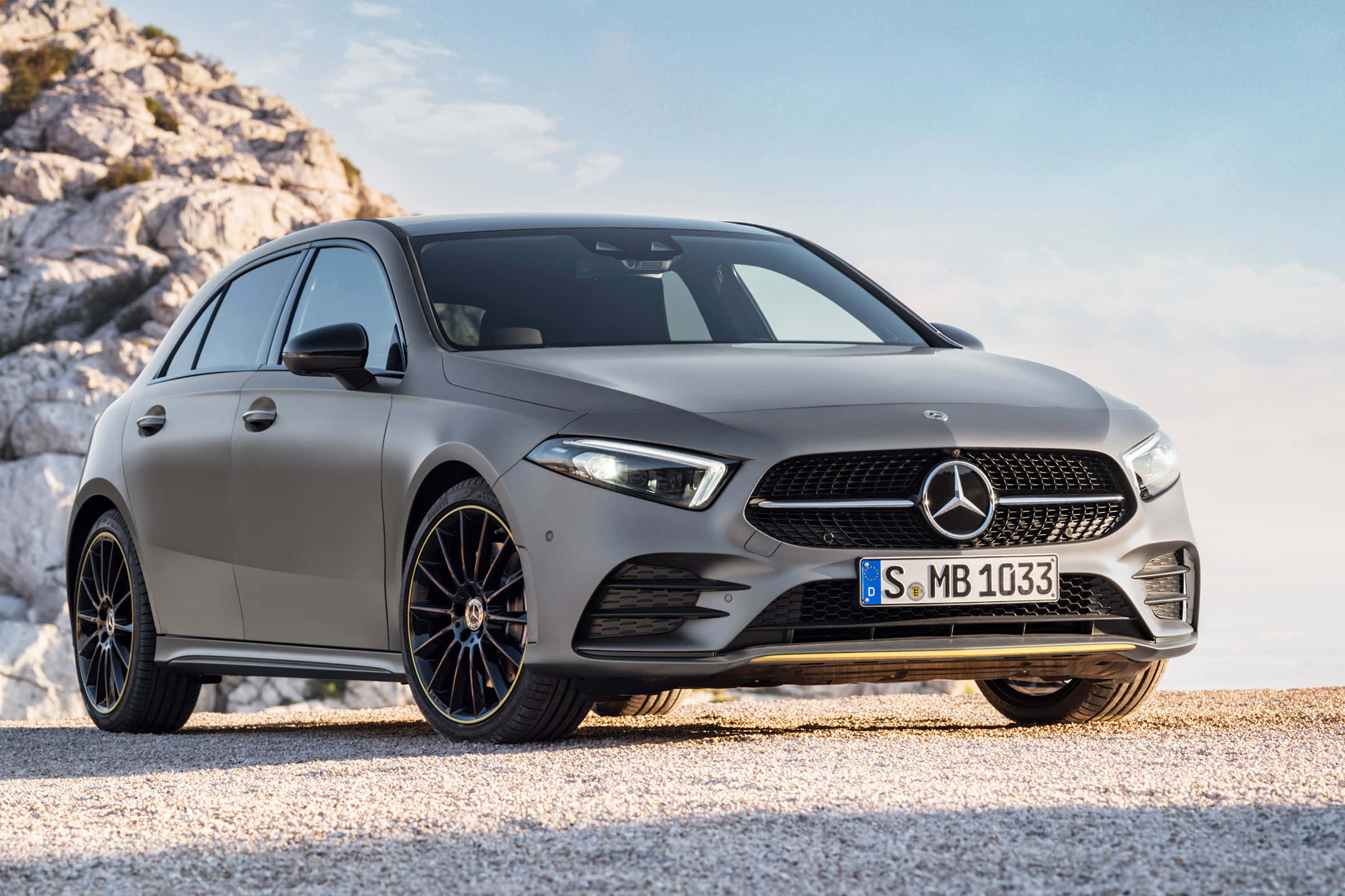 Mercedes Benz A-class 桌布 壁紙 — 一台時尚的消光灰色賓士A-Class停在礫石上，旁邊是岩石峭壁，上方是晴朗的藍天。
