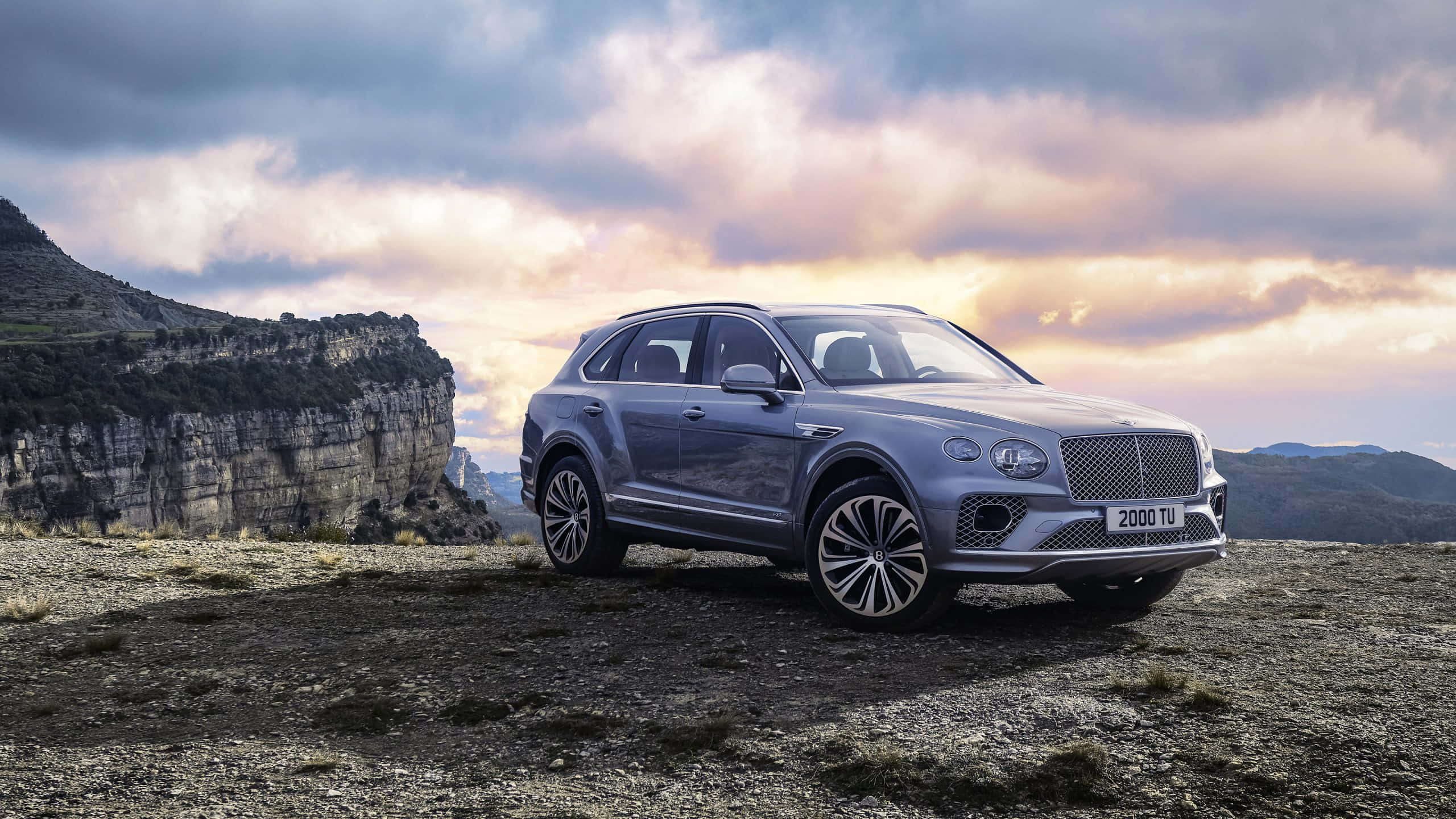 Bentley Bentayga 桌布 壁紙 — 一台時尚的銀色賓利Bentayga SUV在日落時分矗立在崎嶇的懸崖上，背後是戲劇性的雲層和岩石地形。