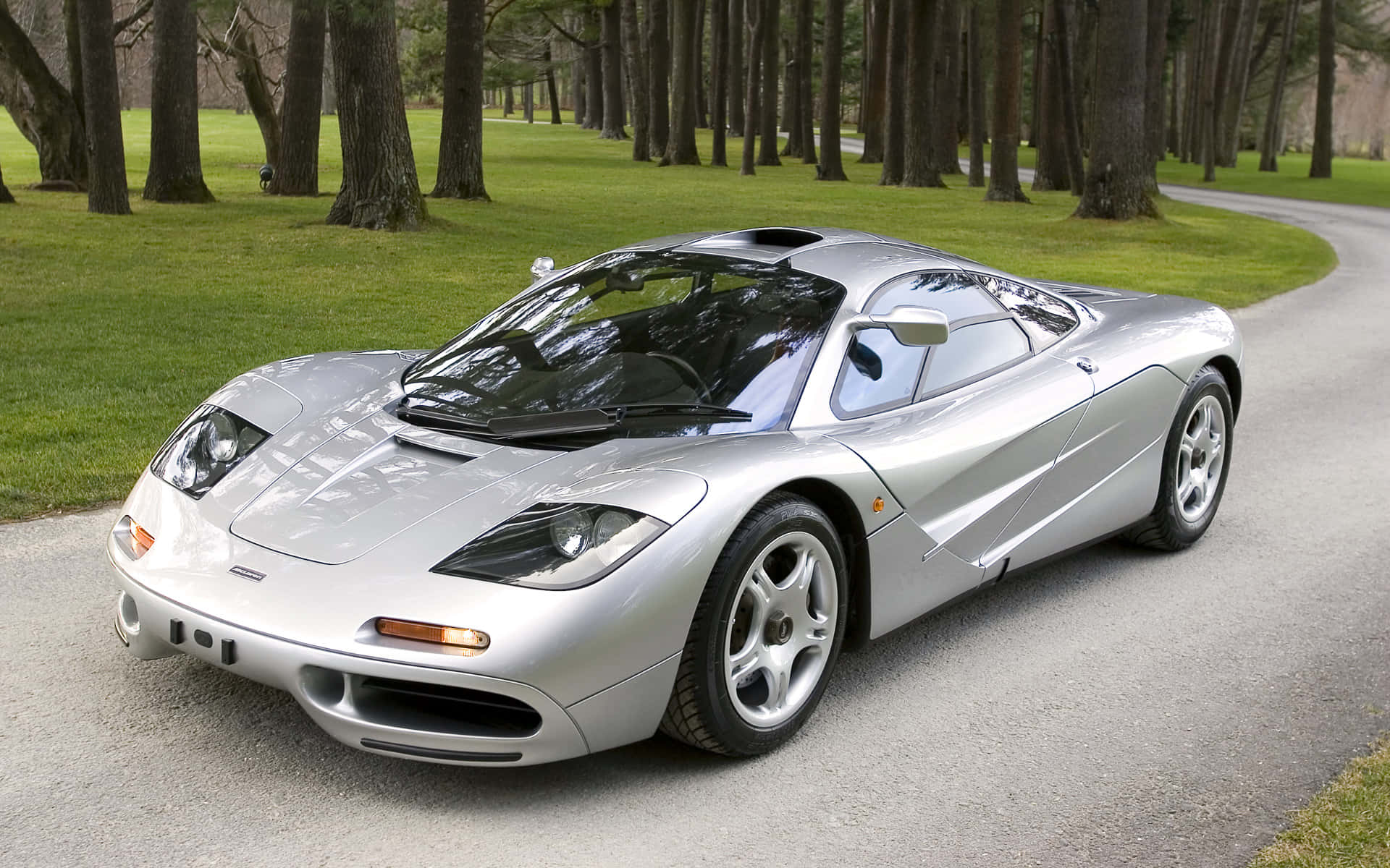 Mclaren F1 桌布 壁紙 — 一台時尚的銀色McLaren F1超級跑車停放在蜿蜒的林蔭大道上，周圍有茂密的綠草。
