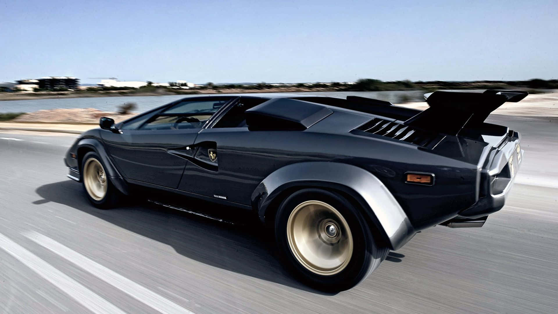 Lamborghini Countach 桌布 壁紙 — 一台時尚的黑色藍寶堅尼Countach在晴朗藍天下沿著海岸高速公路奔馳。
