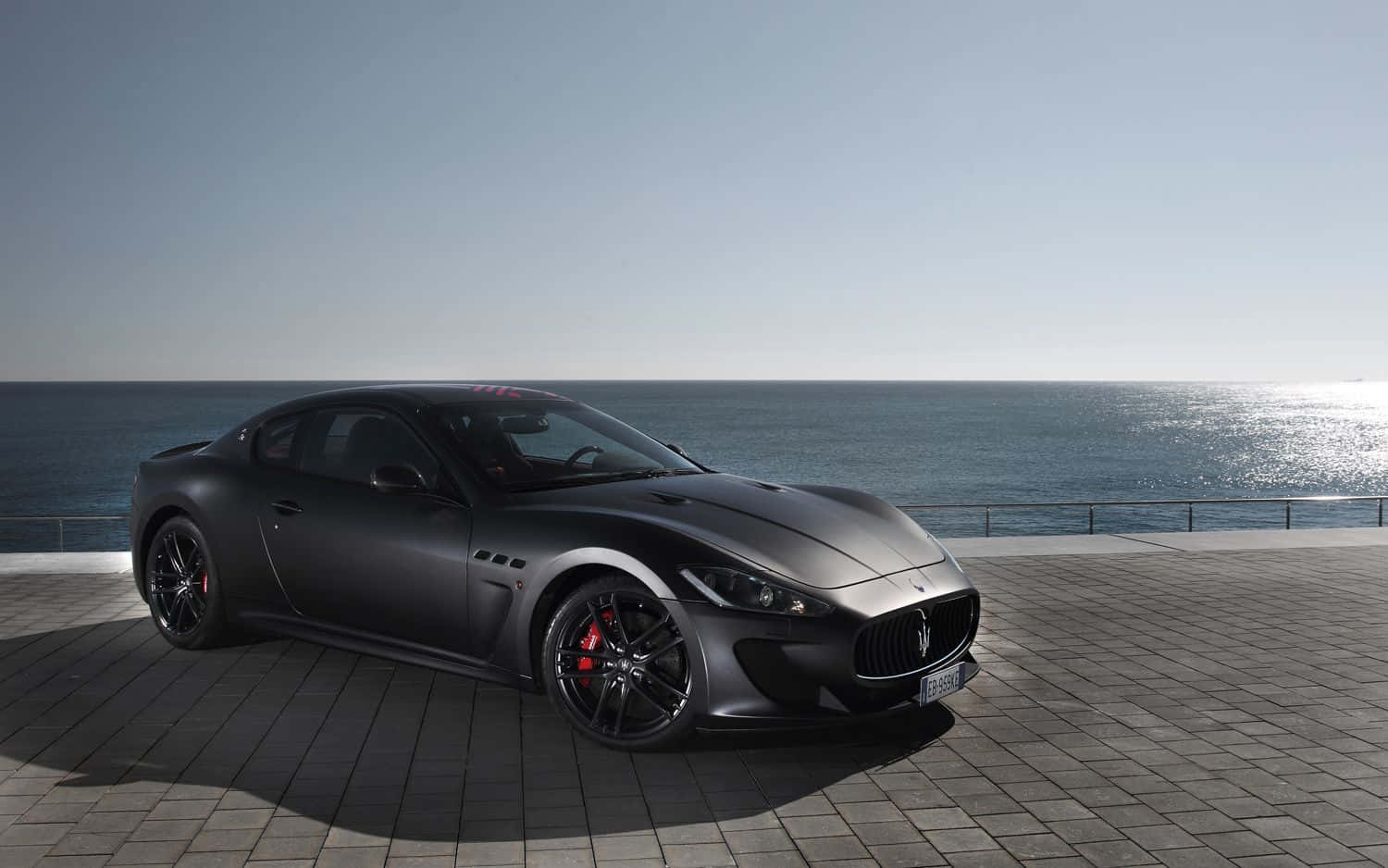 Maserati Granturismo 桌布 壁紙 — 一台時尚的啞光灰色瑪莎拉蒂跑車停泊在磁磚鋪設的海濱長廊上，海平線在晴朗天空下清晰可見。