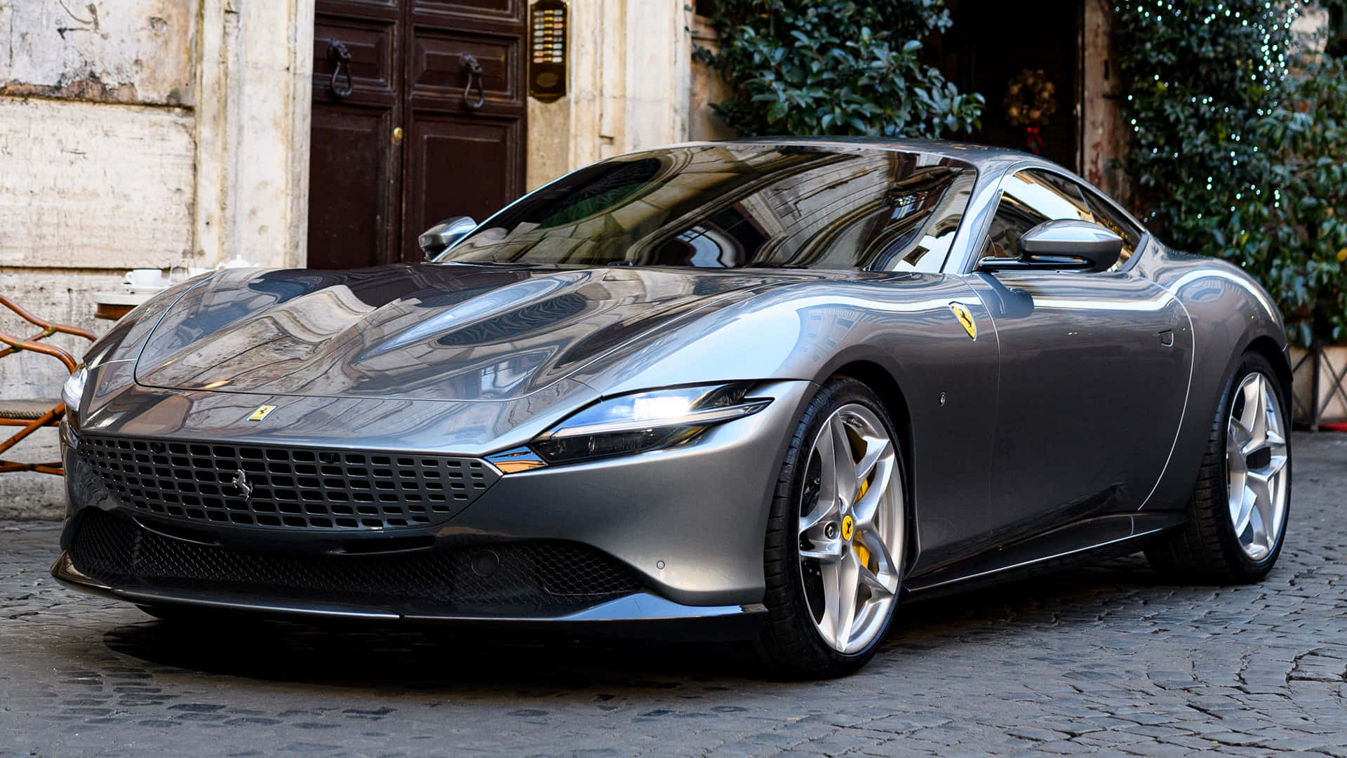 Ferrari Roma 桌布 壁紙 — 一台時尚的銀色法拉利停泊在鵝卵石街道上，旁邊是帶有木門和綠植的歷史建築。