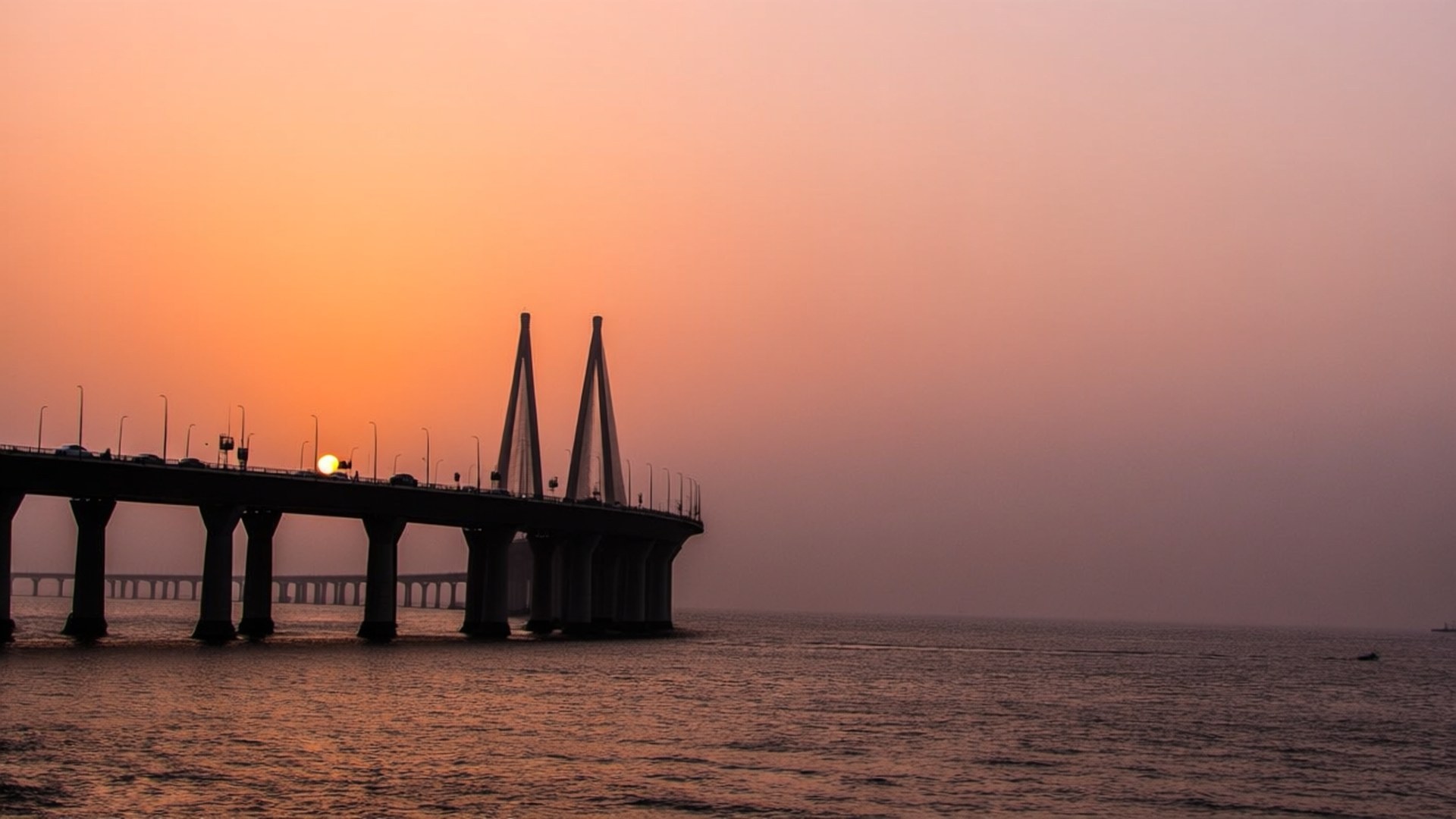 Sea Link Sunset 壁紙 — 一條長長的吊橋在平靜的水面上，襯托著發光的橙色日落，形成剪影。