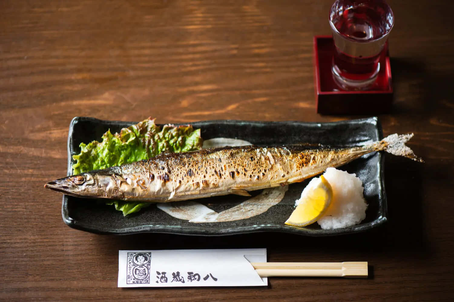  壁紙 — 一條烤全鯖魚放在黑色盤子上，配上米飯、檸檬和生菜，置於木桌上，附有筷子及一杯紅色飲料。