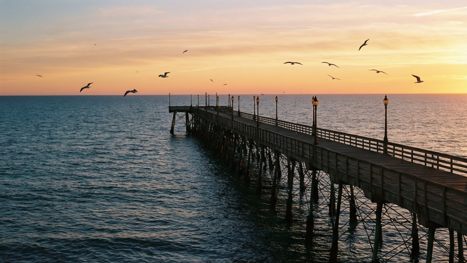 Pier Friday Sunset 壁紙 — 一條木製碼頭在日落時延伸至平靜的海水，上空有剪影鳥兒飛翔。
