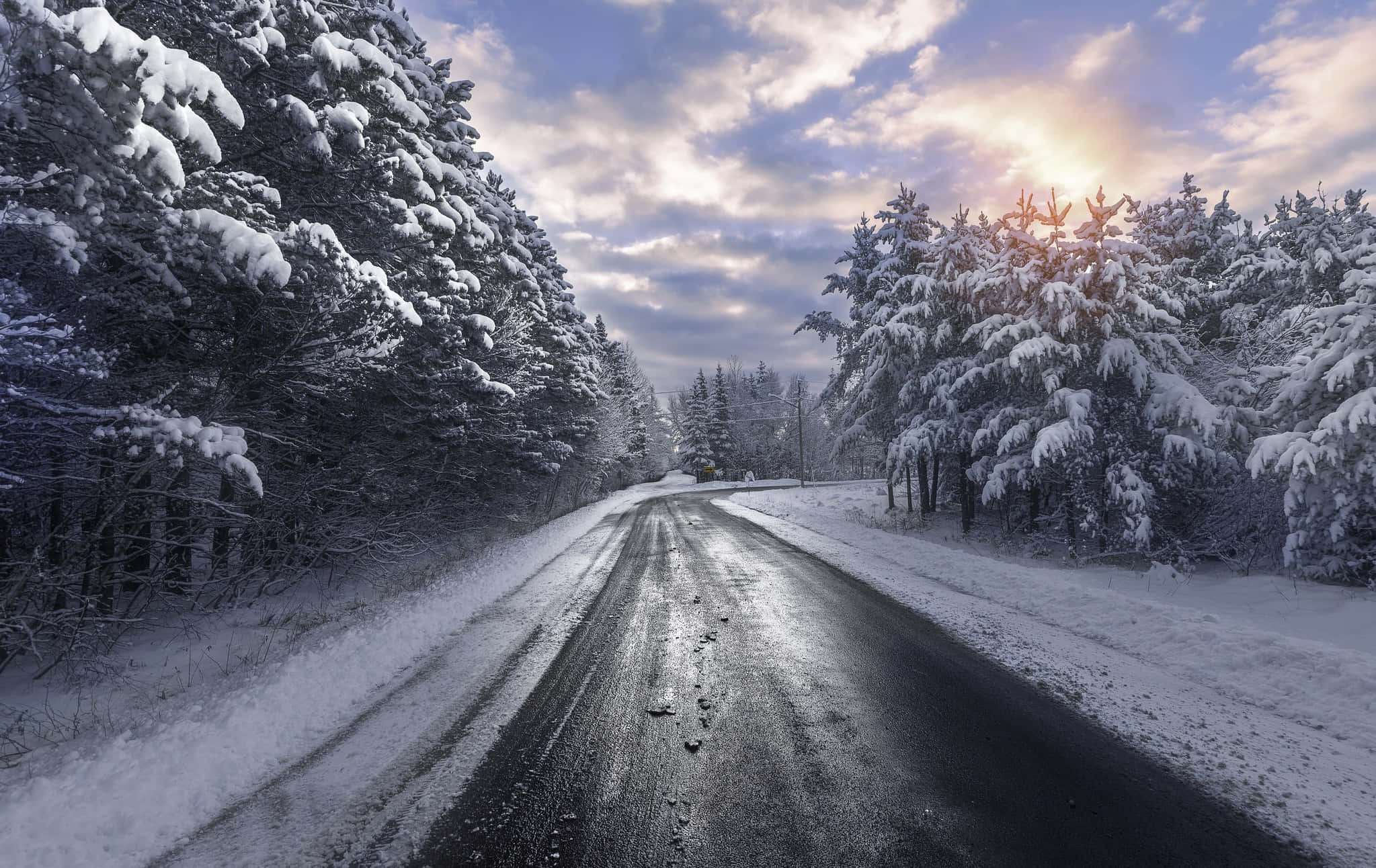 Snowy Road 桌布 壁紙 — 一條蜿蜒的道路穿過白雪覆蓋的森林，太陽從樹後升起，在雲層上投下溫暖的光芒。